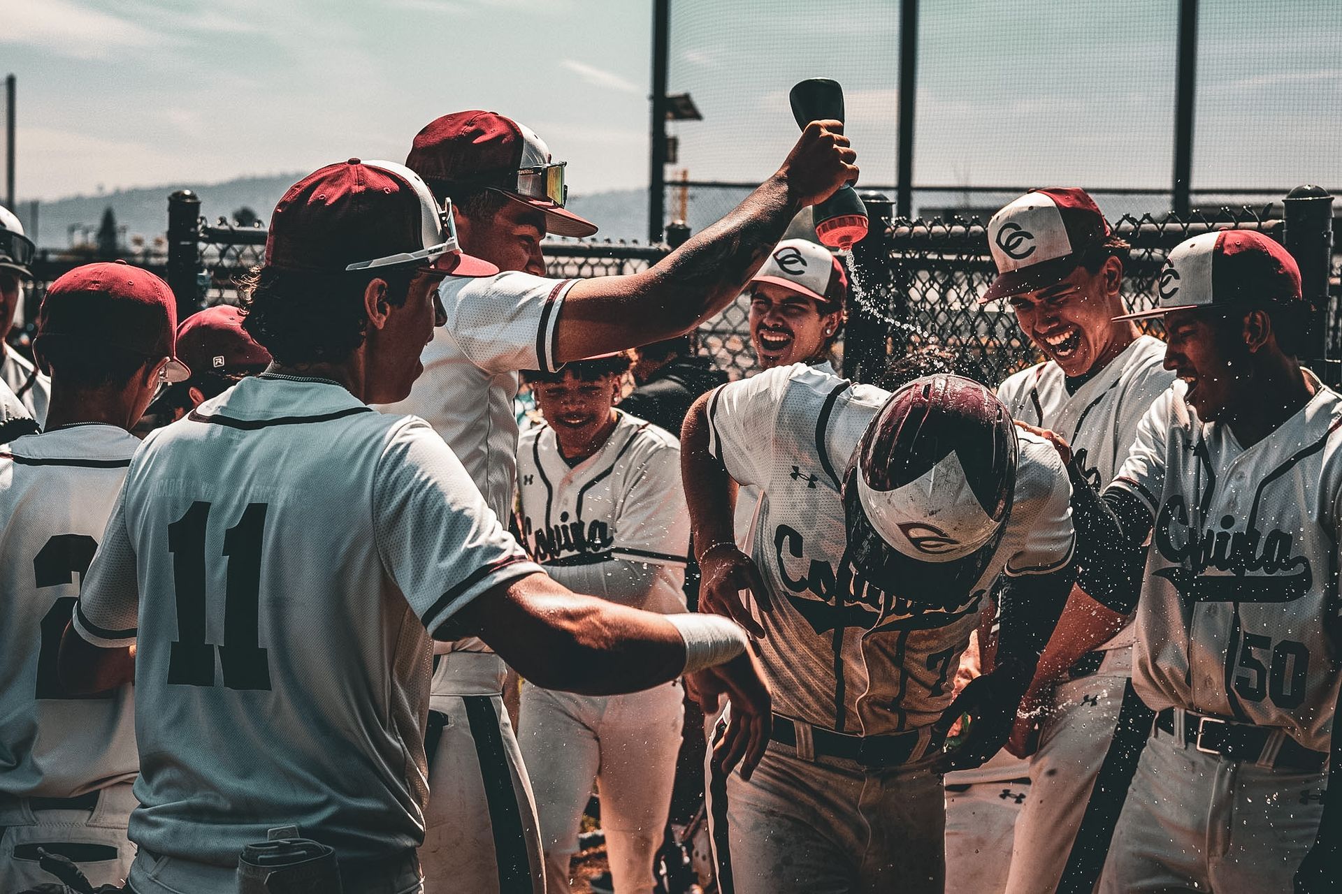 Baseball players in white uniforms and red caps huddle on the field, raising arms in celebration.
