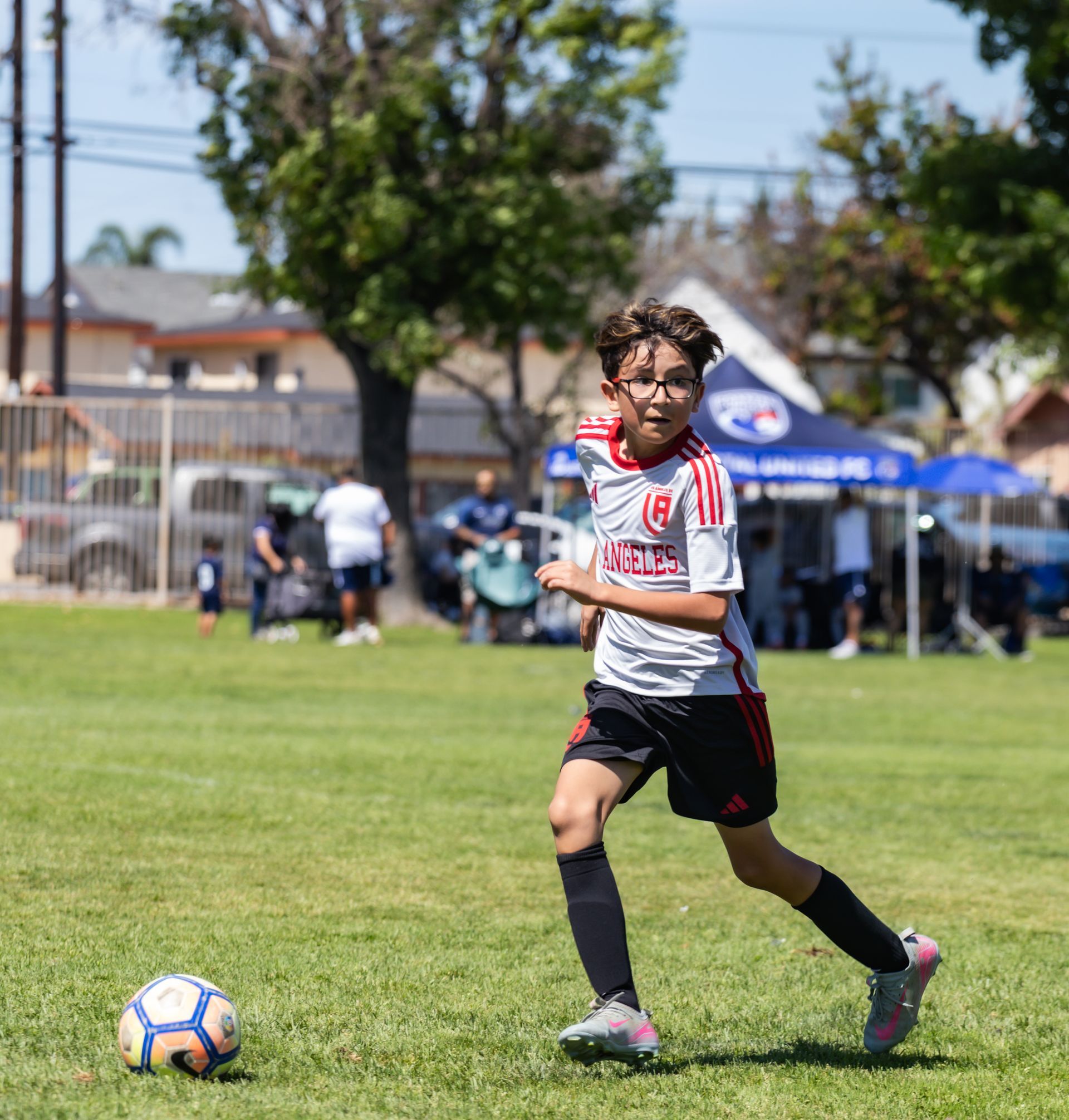 Soccer player in a white jersey dribbles a ball on a grassy field during a sunny match.