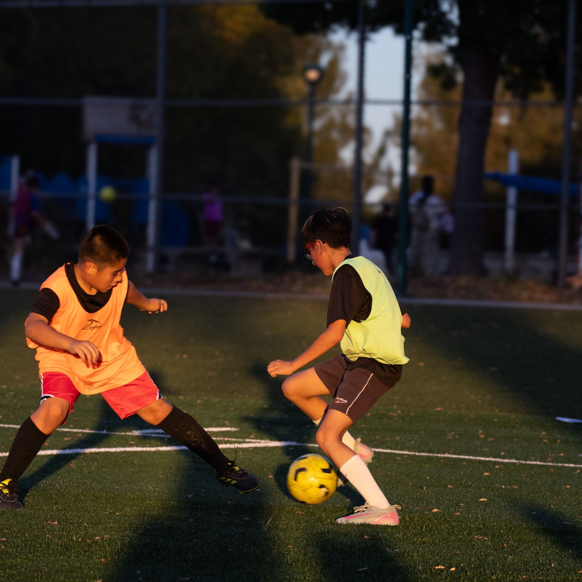 Two soccer players in orange and yellow vests contest a ball on a lit field at dusk