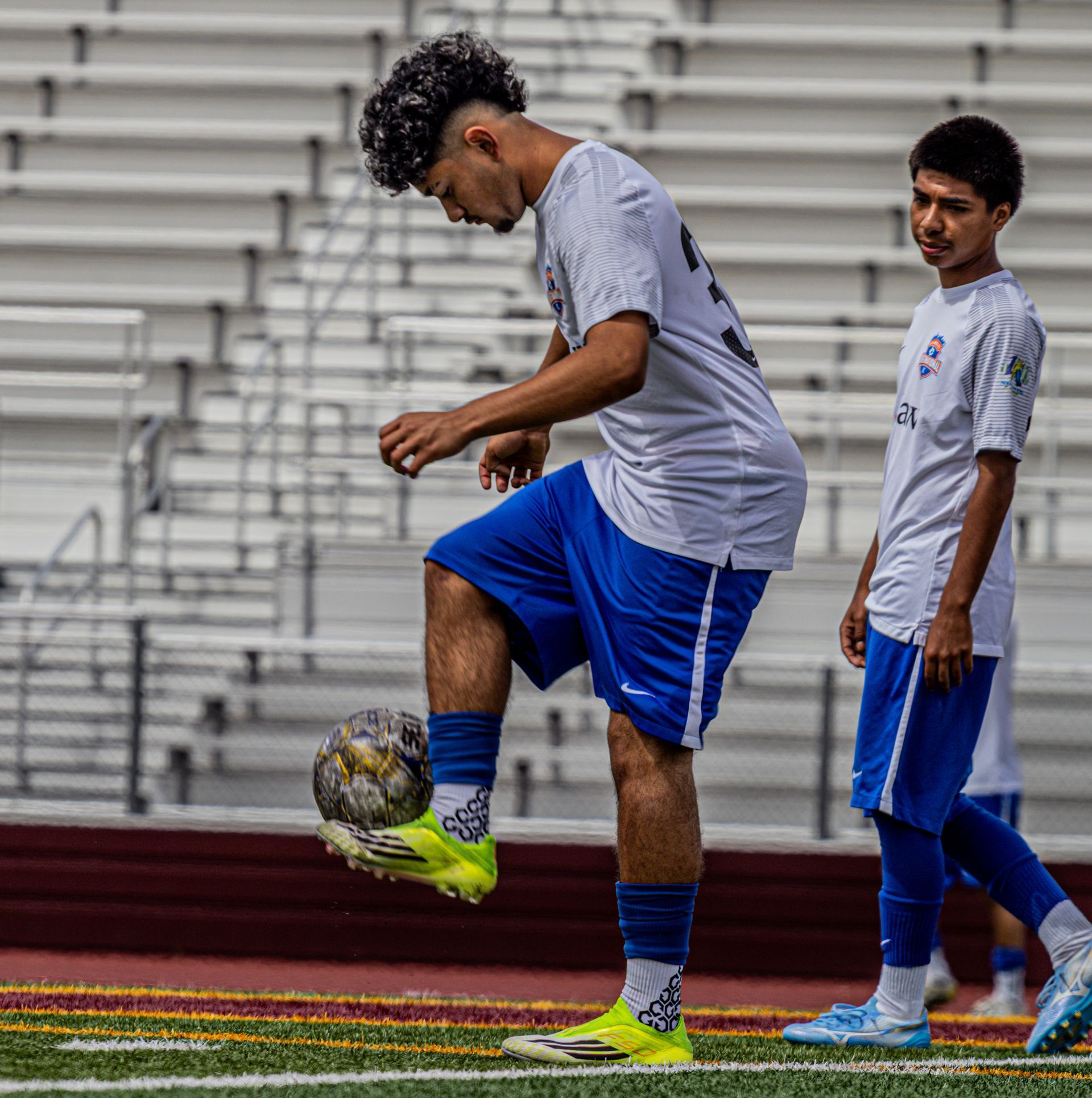 Two soccer players in gray and blue uniforms practice on a field, one juggling a ball.