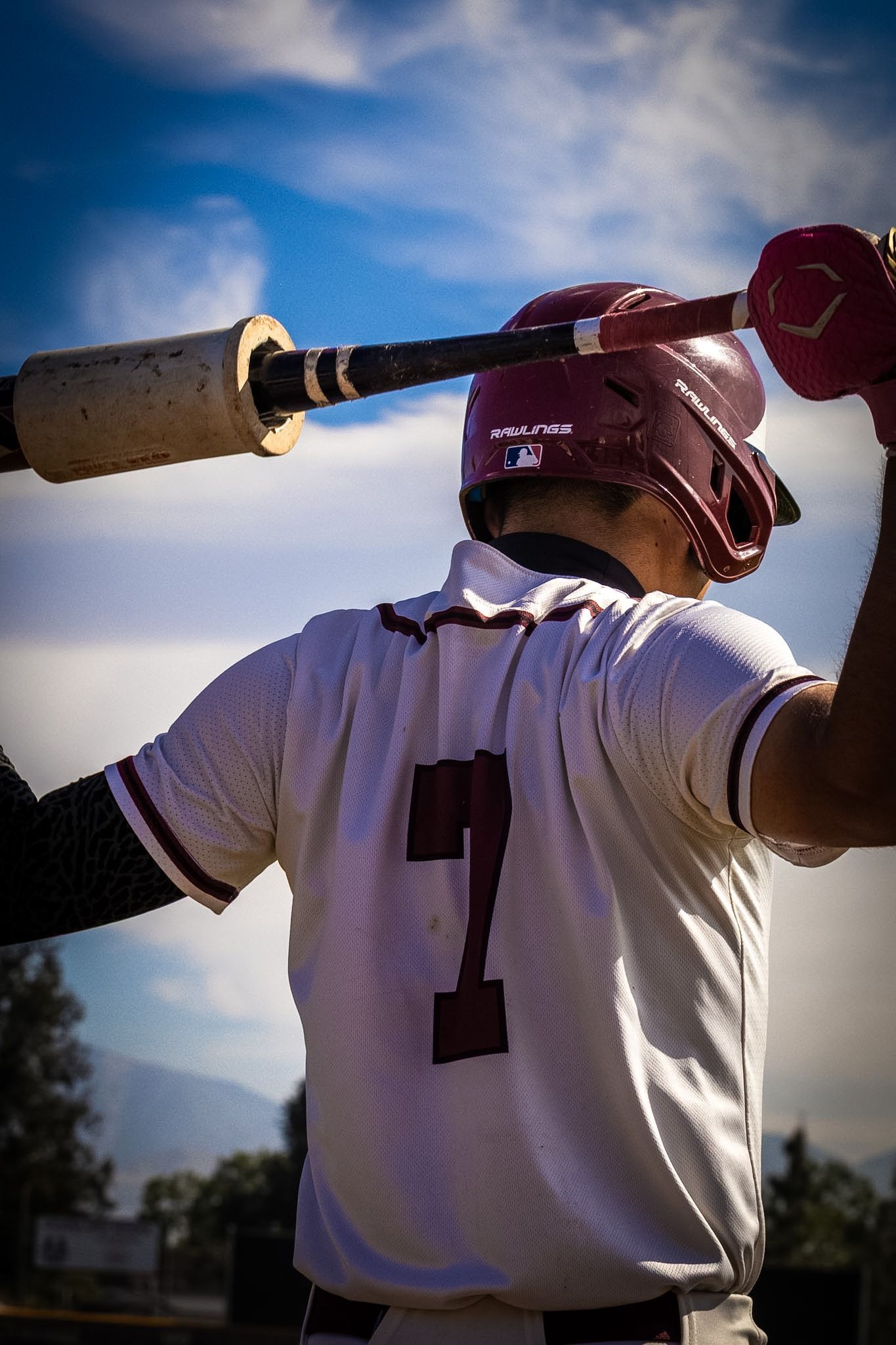 Baseball player in maroon jersey holding a bat on his shoulder against a blue sky