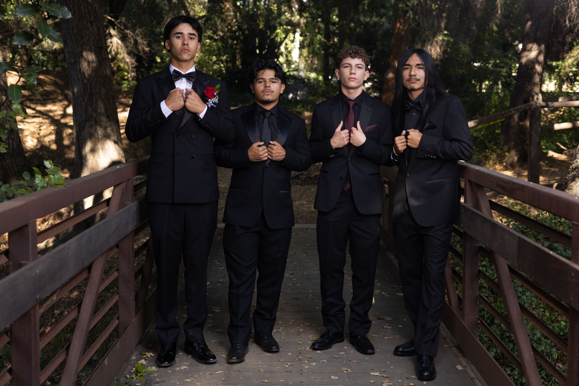 Four people in black suits stand on a wooden bridge in a wooded outdoor setting.