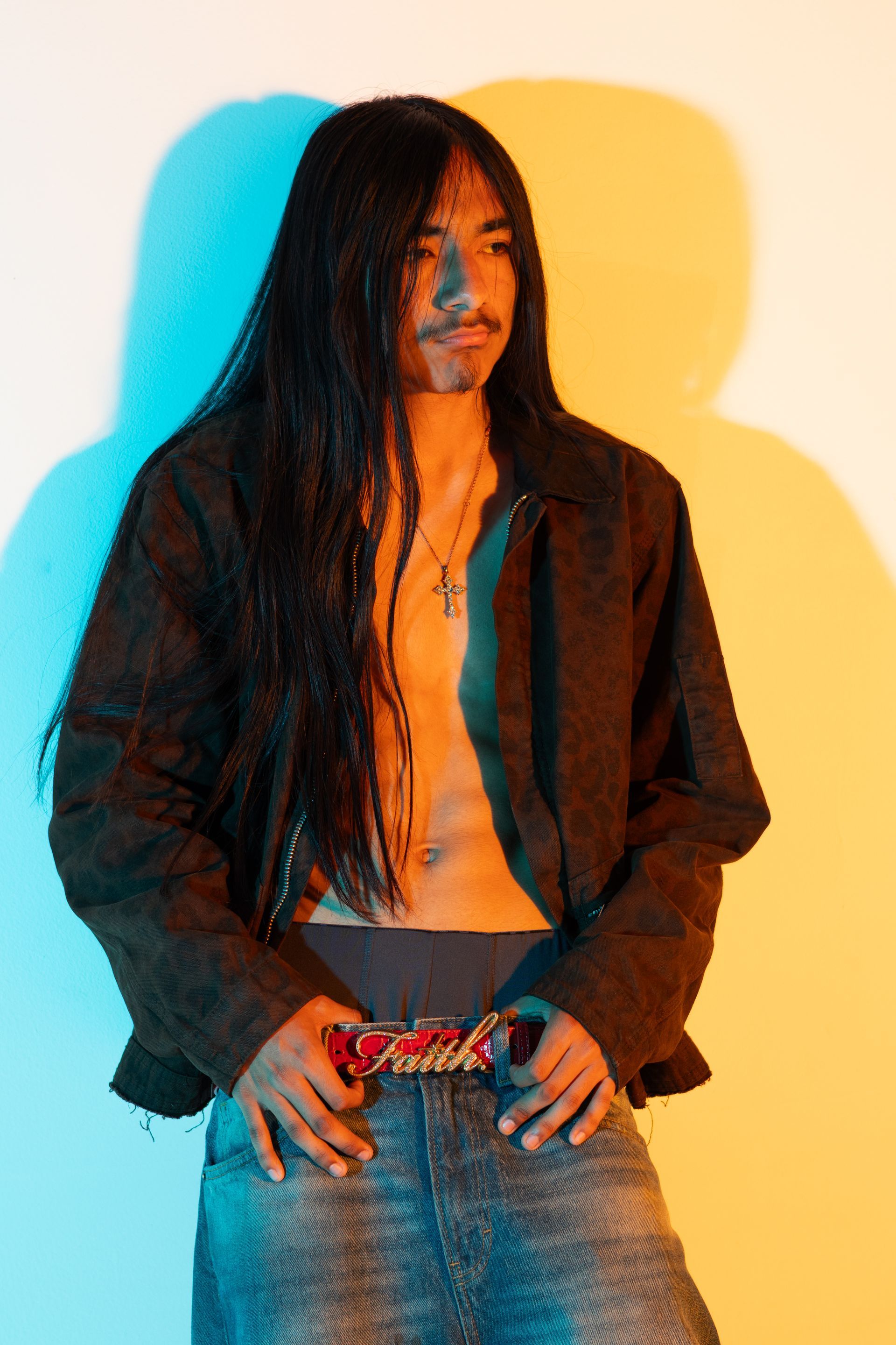 Person with long black hair in a dark jacket and jeans, standing against a white wall with blue and yellow lighting.