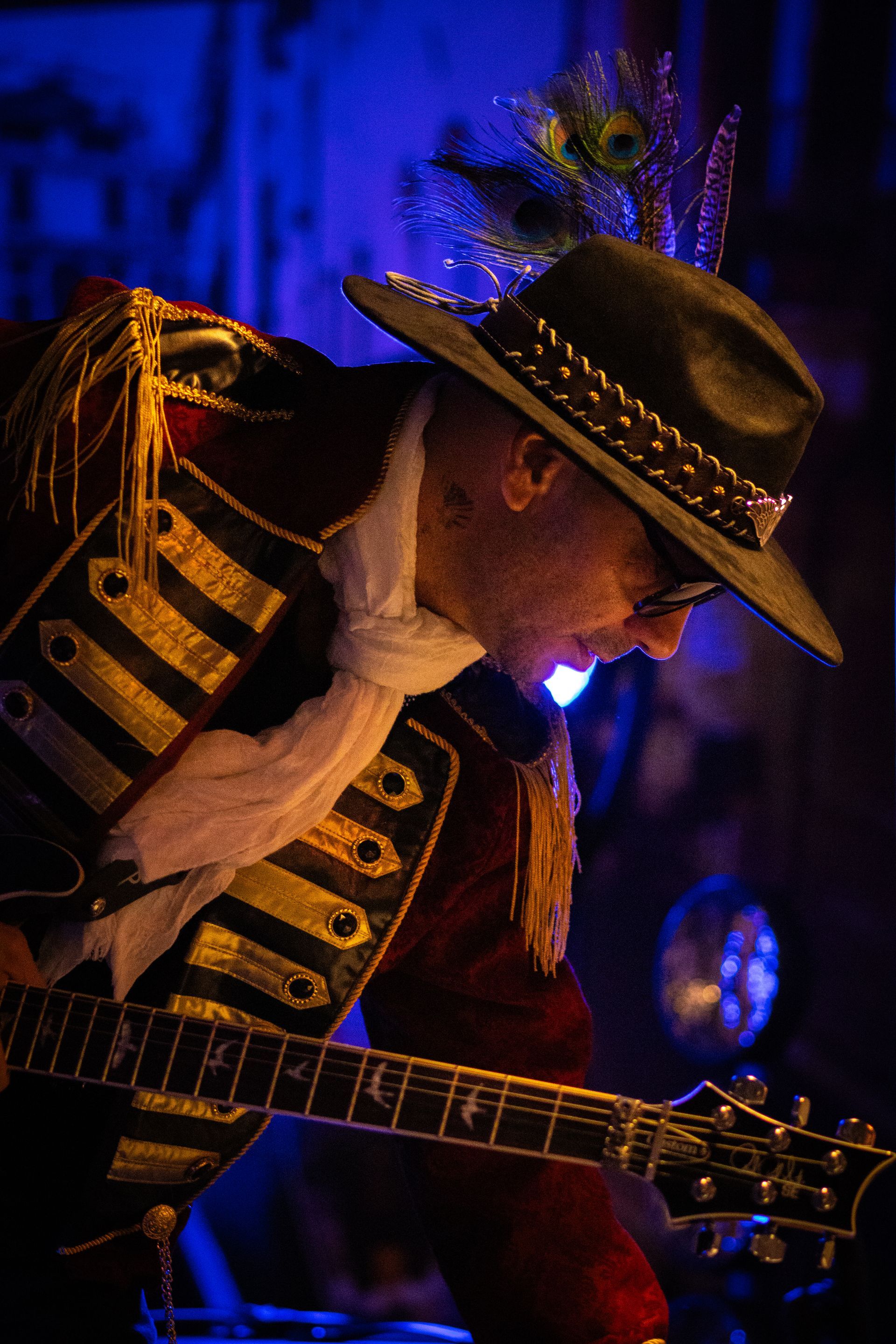 Musician in black-and-gold jacket and hat playing electric guitar under blue stage lighting
