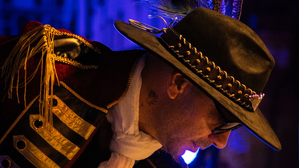 Live concert photography of a musician wearing a decorated black cowboy hat with gold trim and a feather, performing close to the microphone under blue stage lighting.