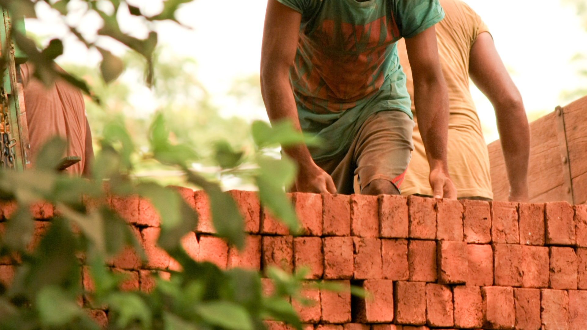 Electrician in orange shirt and overalls working on electrical panel, wearing gloves and yellow hard hat.