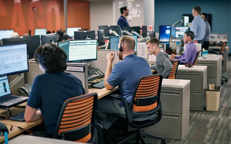 Office with several people at desks, some wearing headsets. Fluorescent lighting.