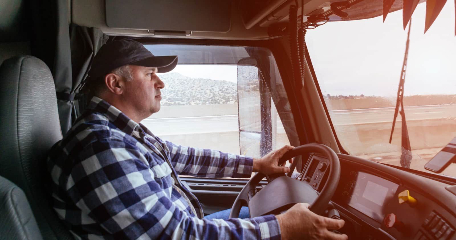 Truck driver in blue plaid shirt and cap, driving a semi-truck; sunny outdoor setting.
