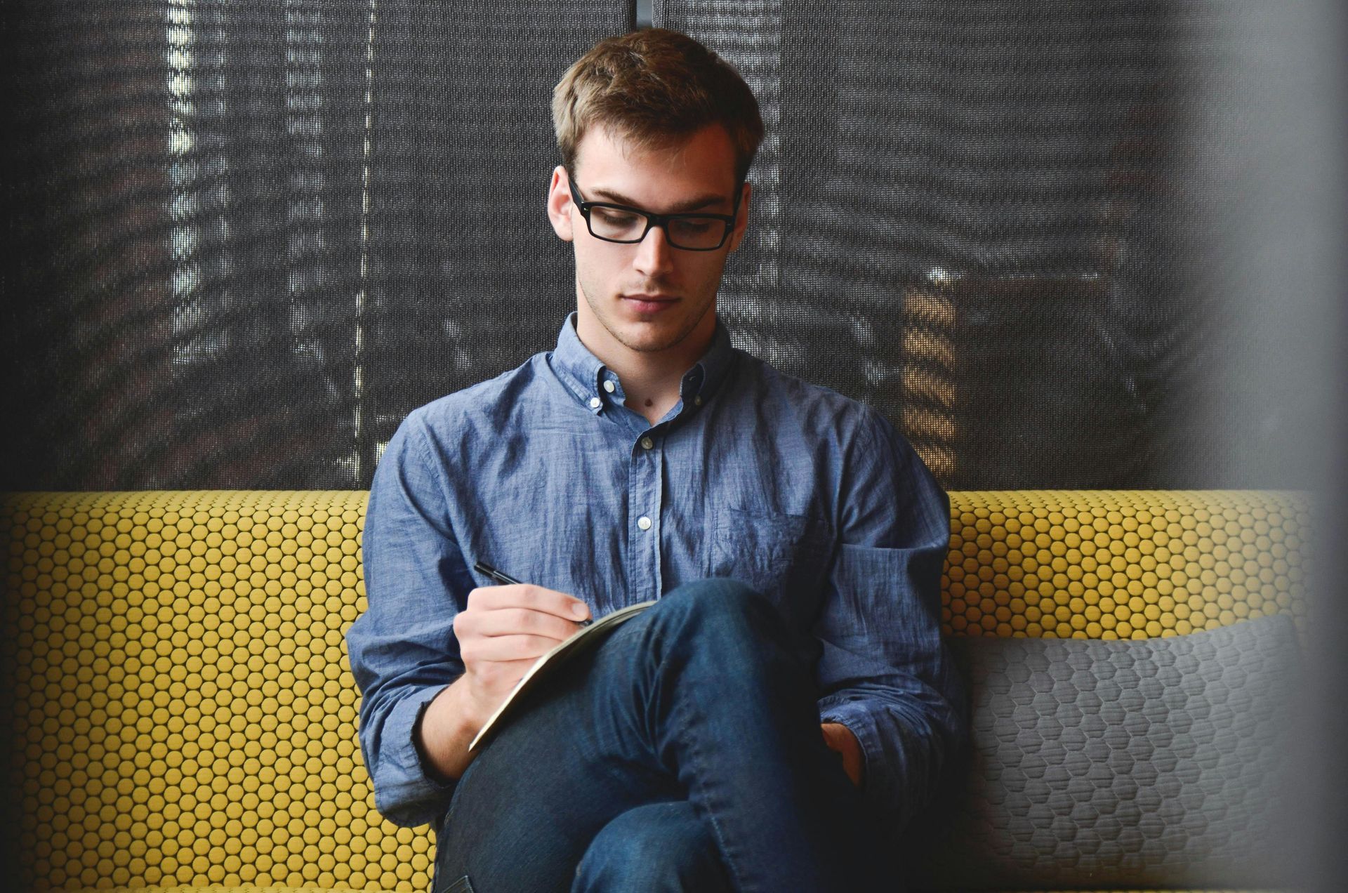 Man in glasses writing in a notebook, seated on a yellow and gray couch indoors.