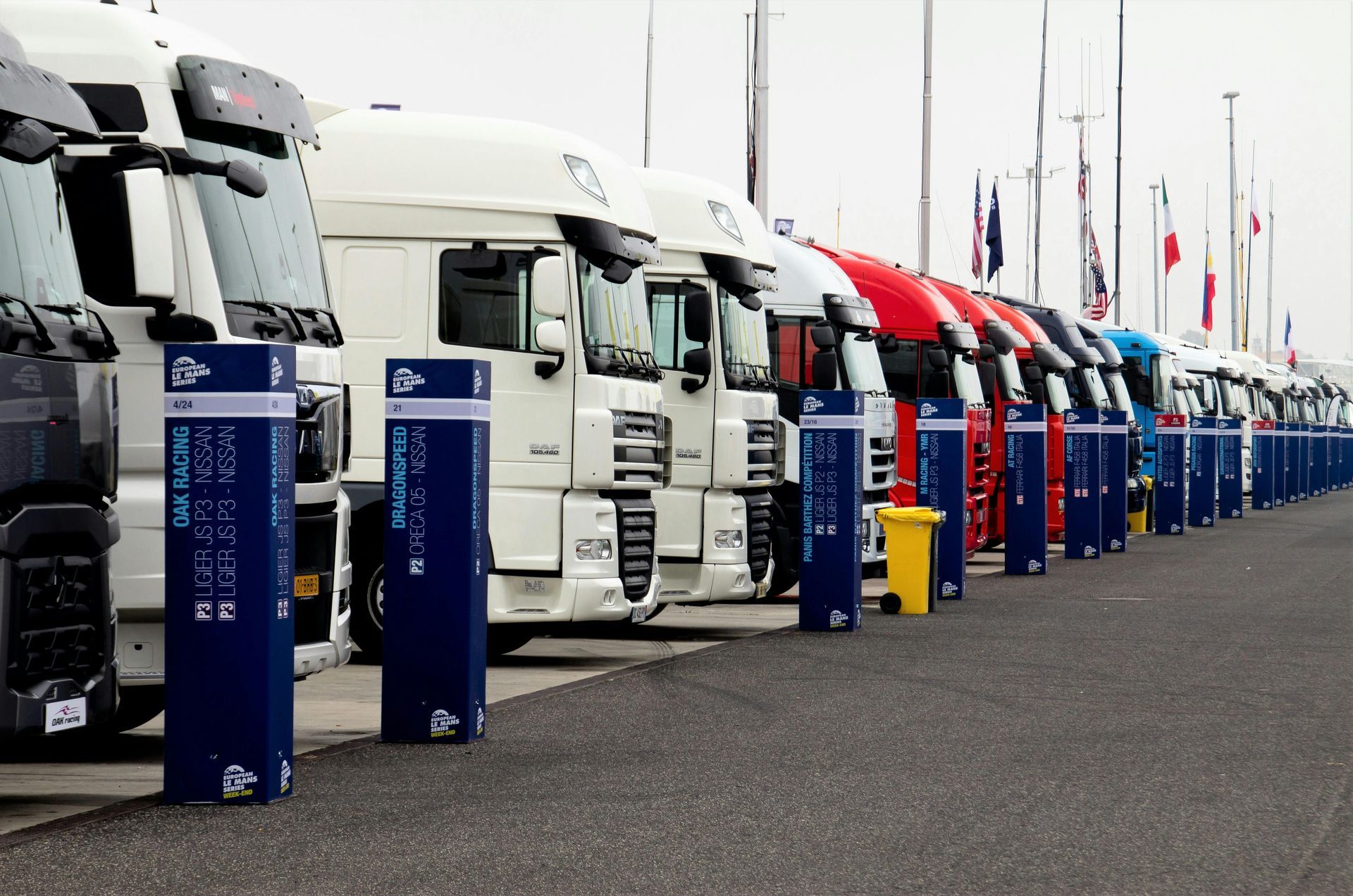Semi-trucks parked in a row, next to blue charging stations. Flags in the background.