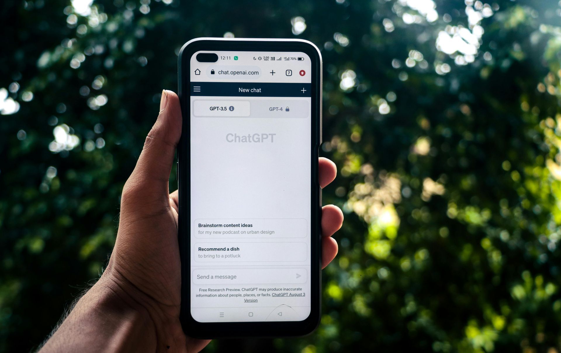 A hand holds a smartphone displaying the ChatGPT website against a blurred, leafy green outdoor background.