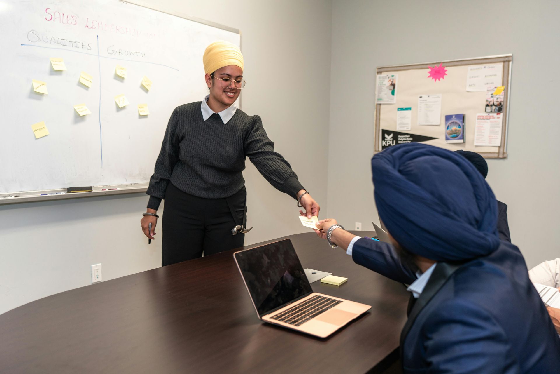 Two people in an office setting engage in a meeting, with one standing at a whiteboard and handing a note to the other.