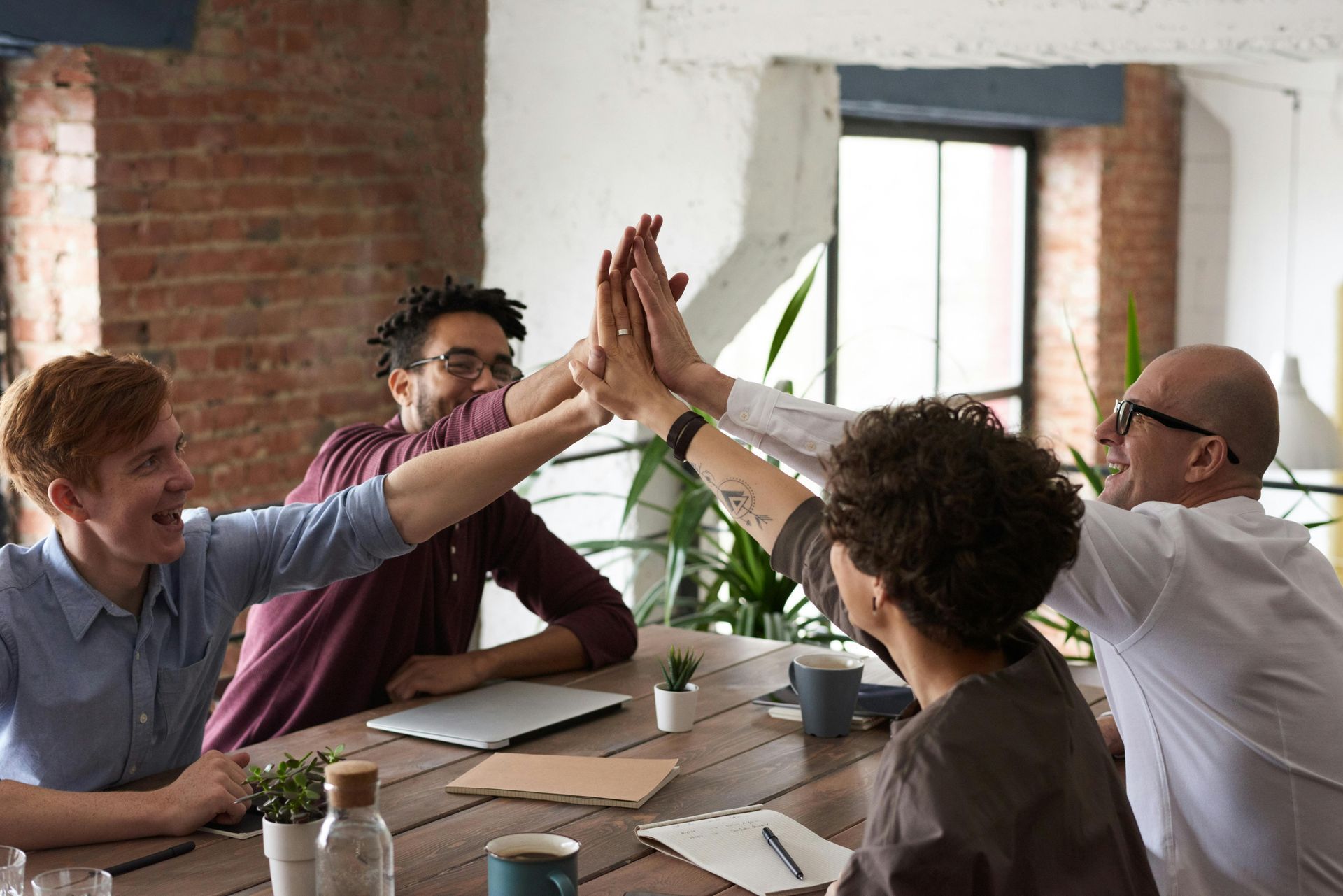 Four people at a table high-fiving in an office setting. They are smiling and appear to be celebrating.