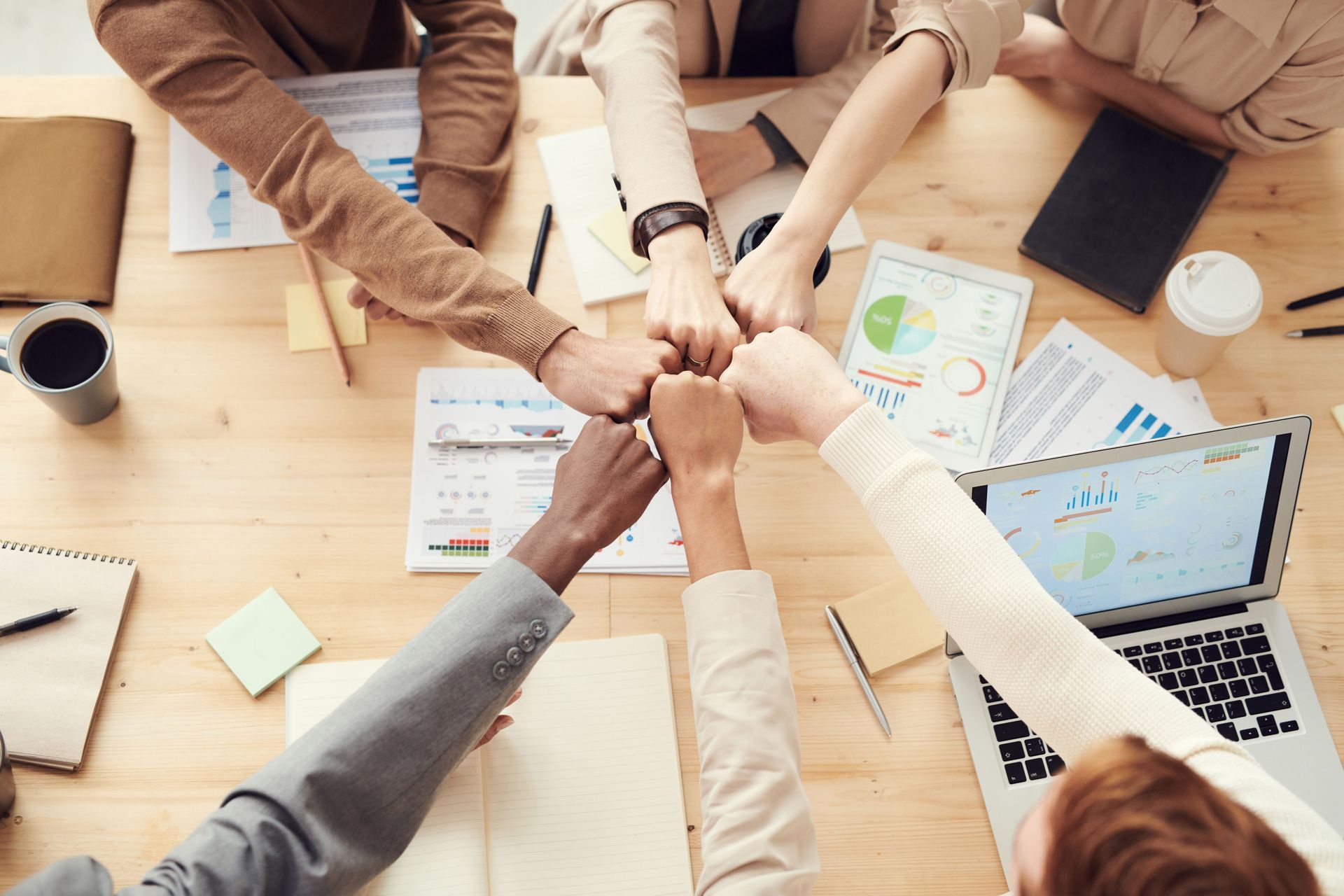 Colleagues stack hands over a desk with charts, coffee, and a laptop in a team meeting