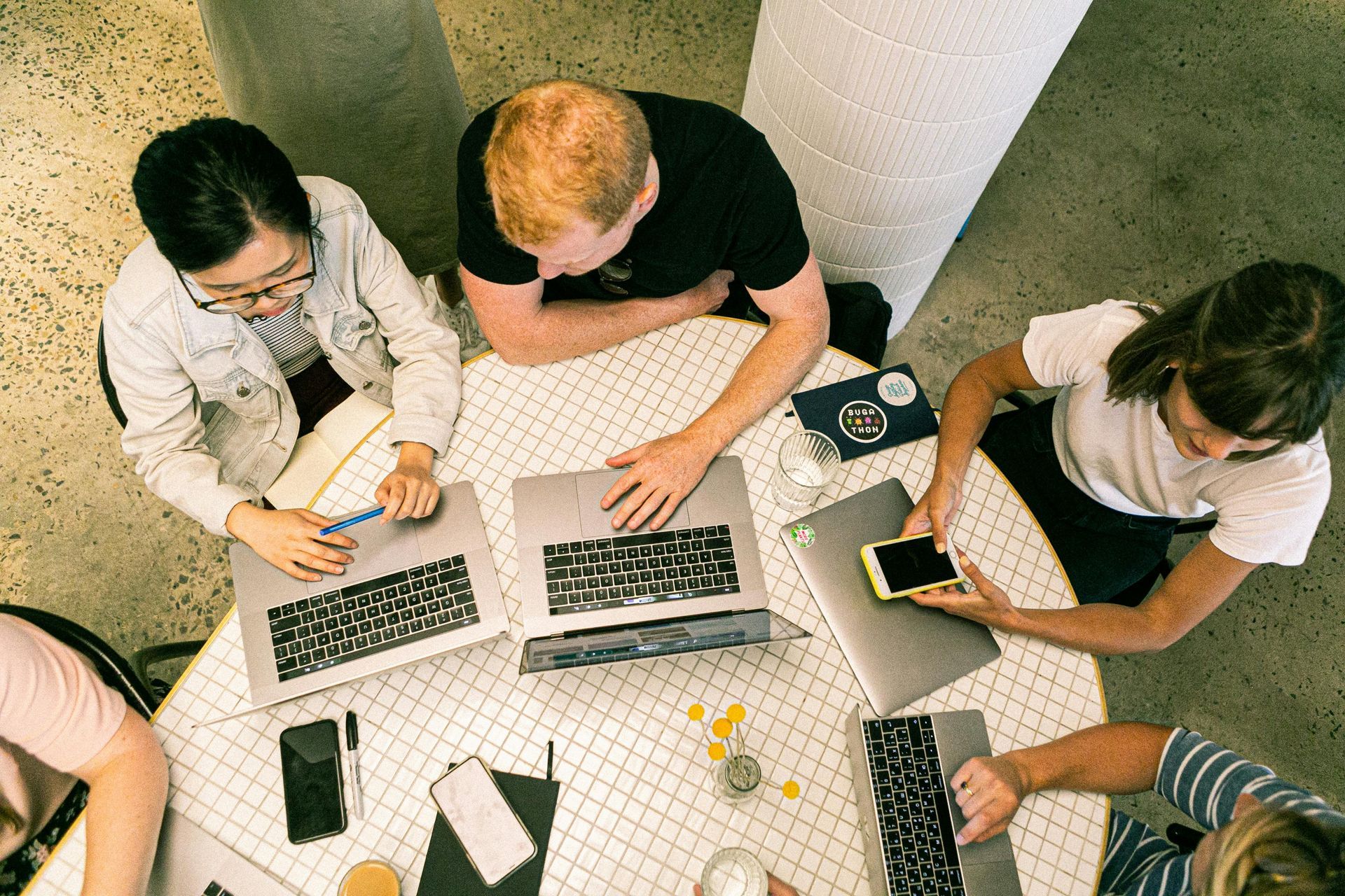 Five people sitting at a circular table in an office setting, working on laptops and using mobile phones.
