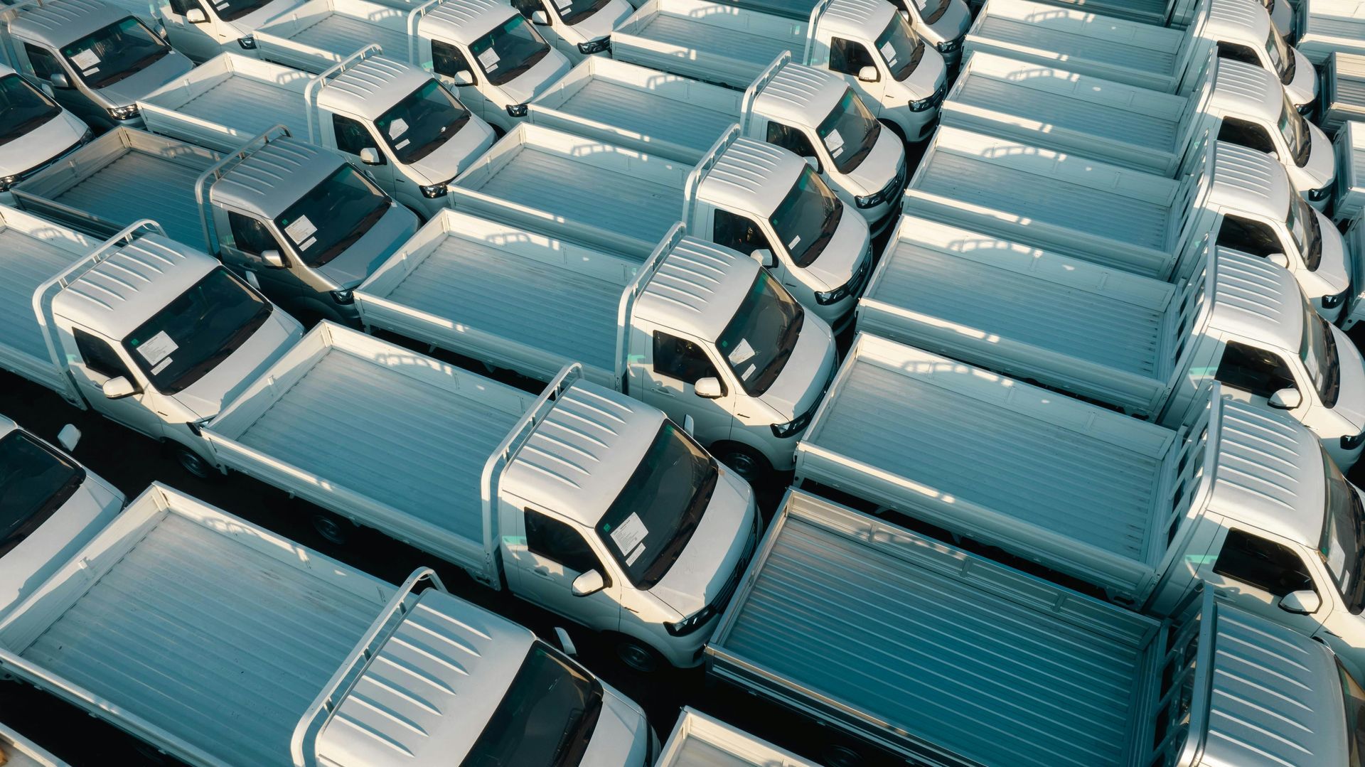 A high-angle view of rows of identical white pickup trucks parked closely together in a large, paved outdoor lot.