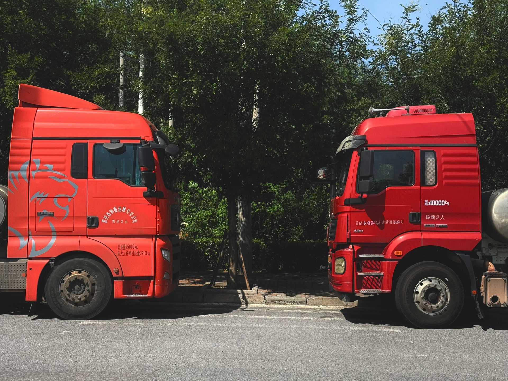 Two red semi-trucks parked side-by-side, facing each other on a road with trees in the background.