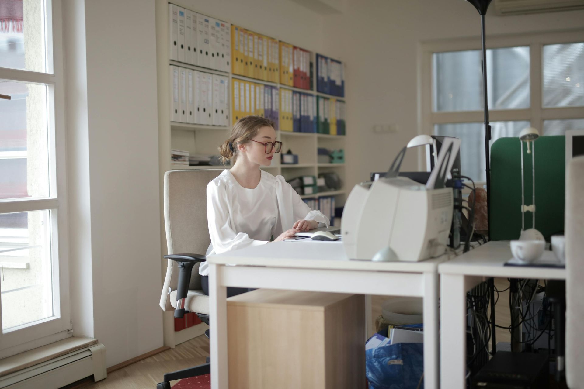 Woman working at a desk in a brightly lit office with files and a printer.