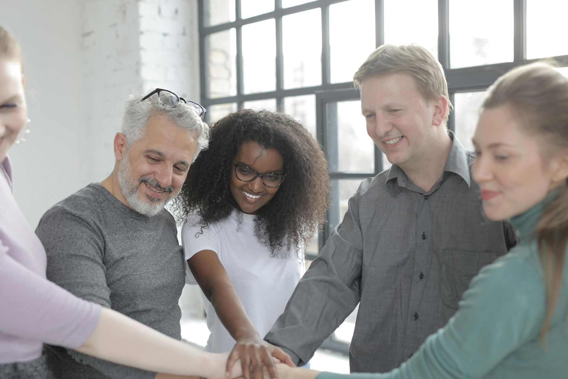 Team of diverse people with hands stacked, smiling, by a window.