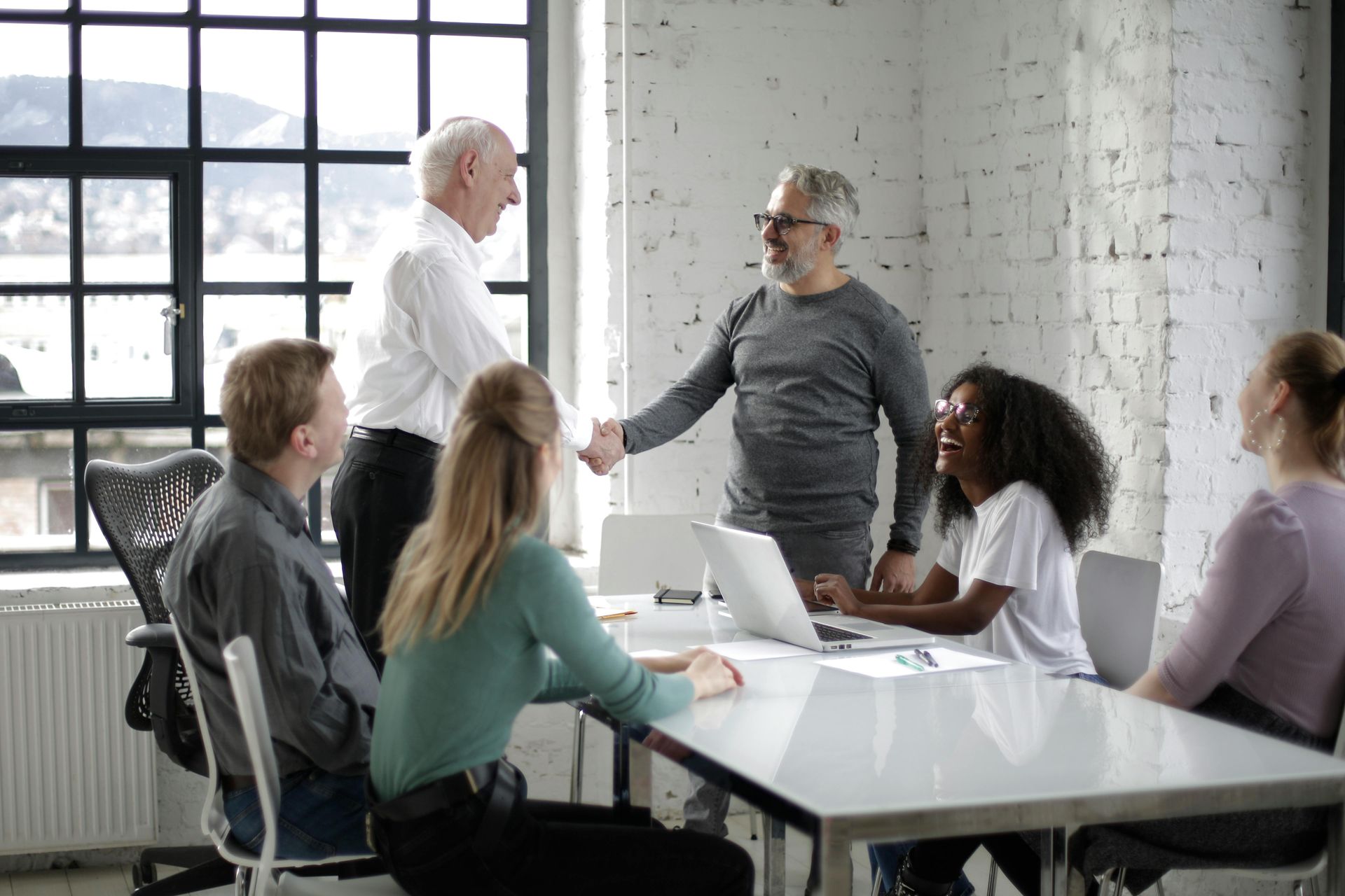 People in an office meeting shaking hands, a laptop on the table, and a window in the background.