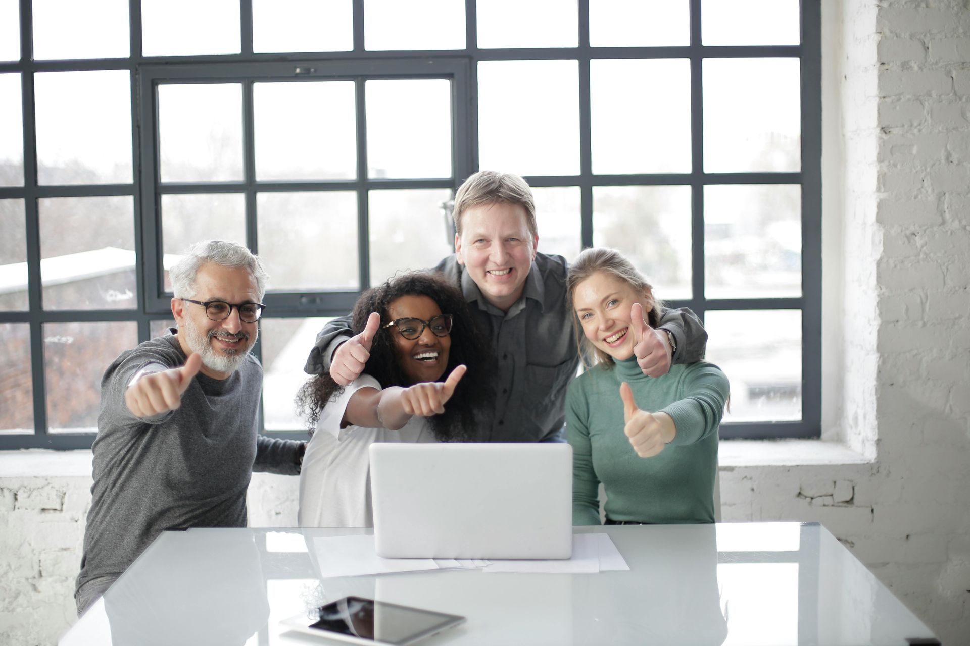 Four people at a table giving thumbs up; laptop in front of them, window in background.