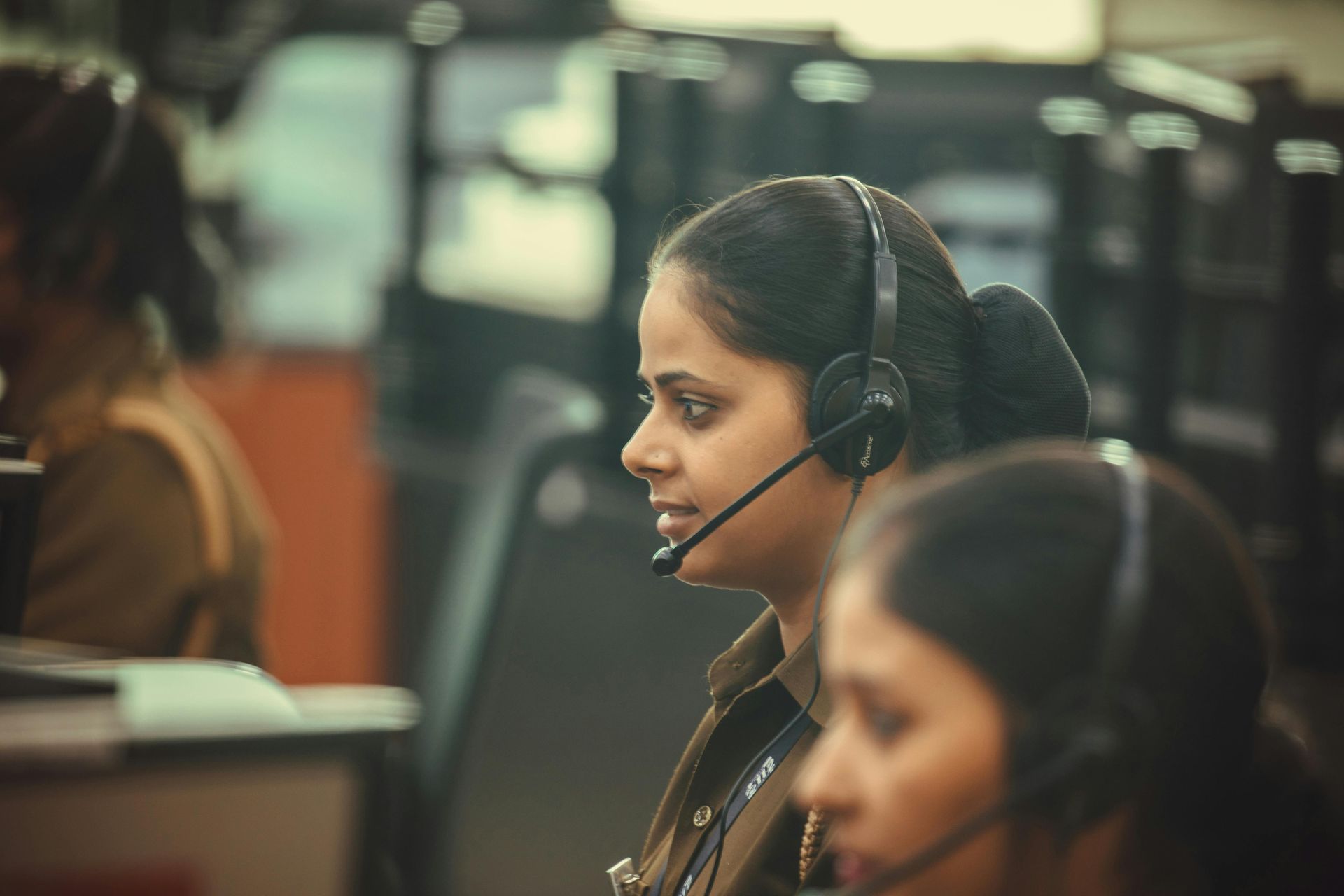 Woman wearing headset at a workstation, focused expression. Others visible. Indoor, office setting.