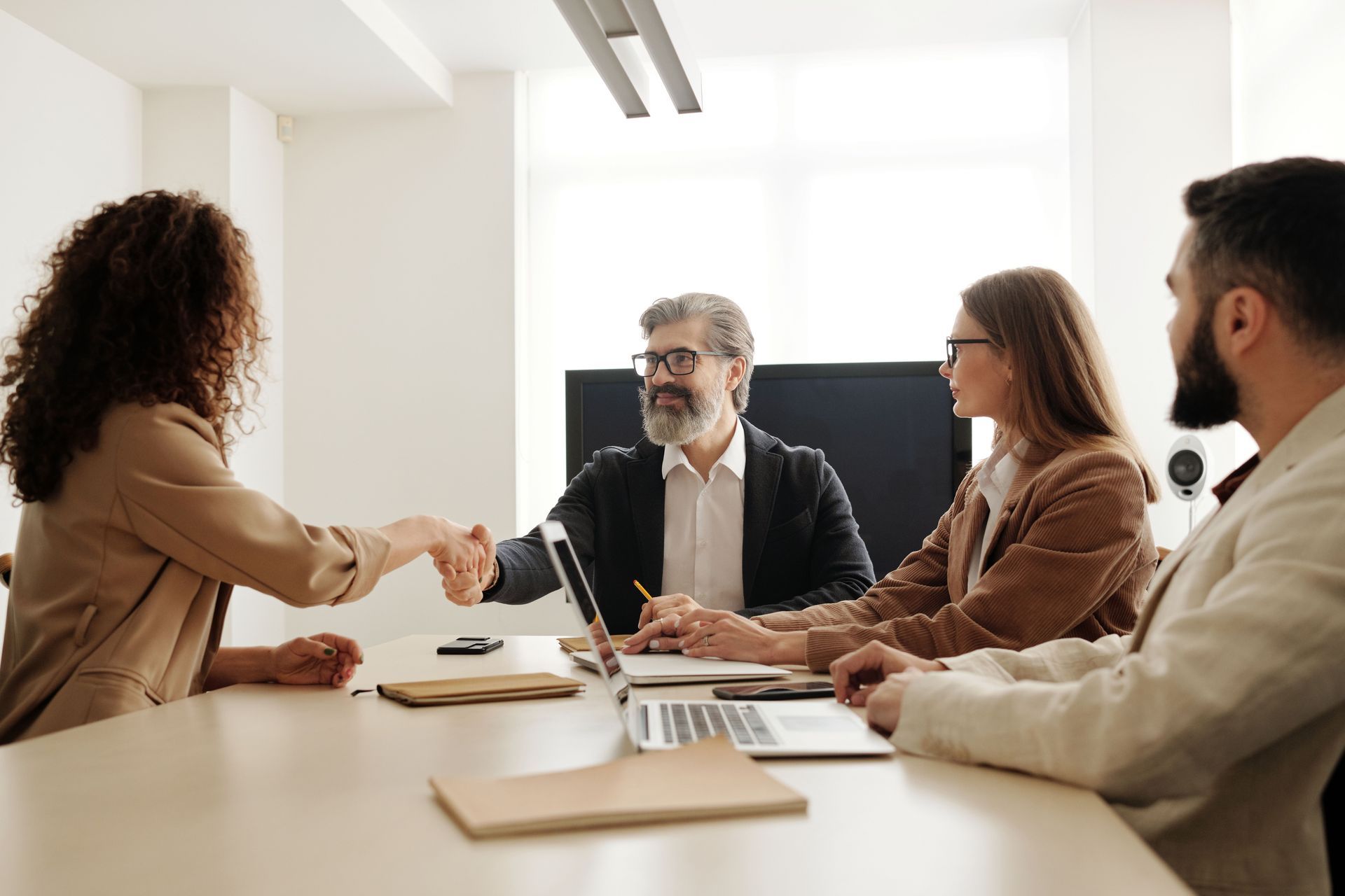 Four colleagues in a bright meeting room shake hands across a conference table.