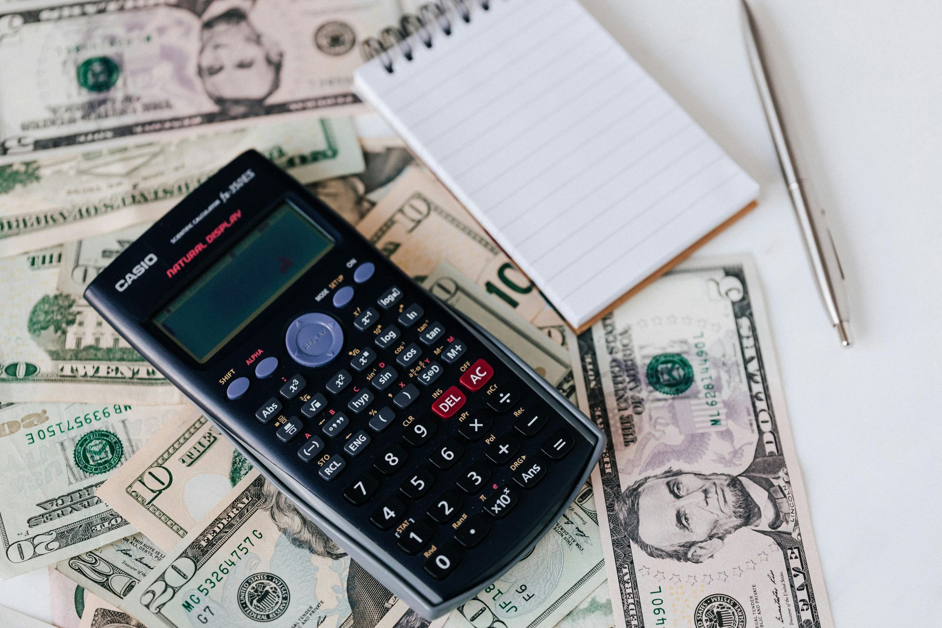 A calculator, a notepad, and a pen resting on a pile of scattered US dollar bills.