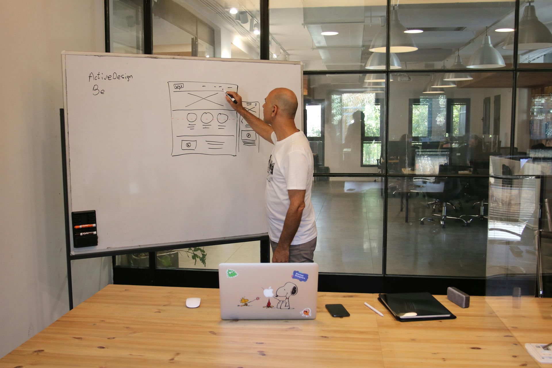 A person writes on a large whiteboard in a bright, modern office with glass walls, facing a table with a laptop.