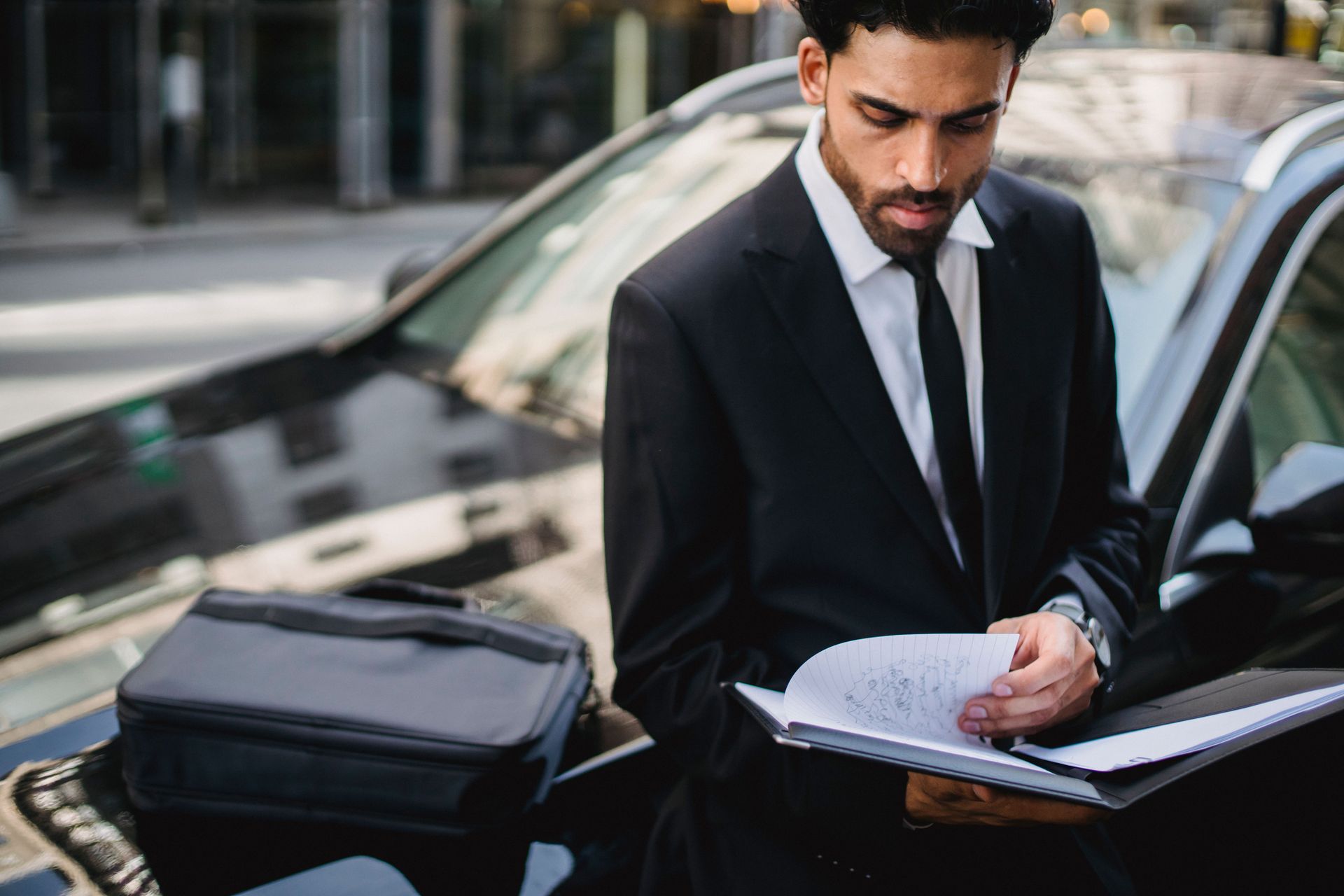 Man in a black suit writing on papers beside an open car door