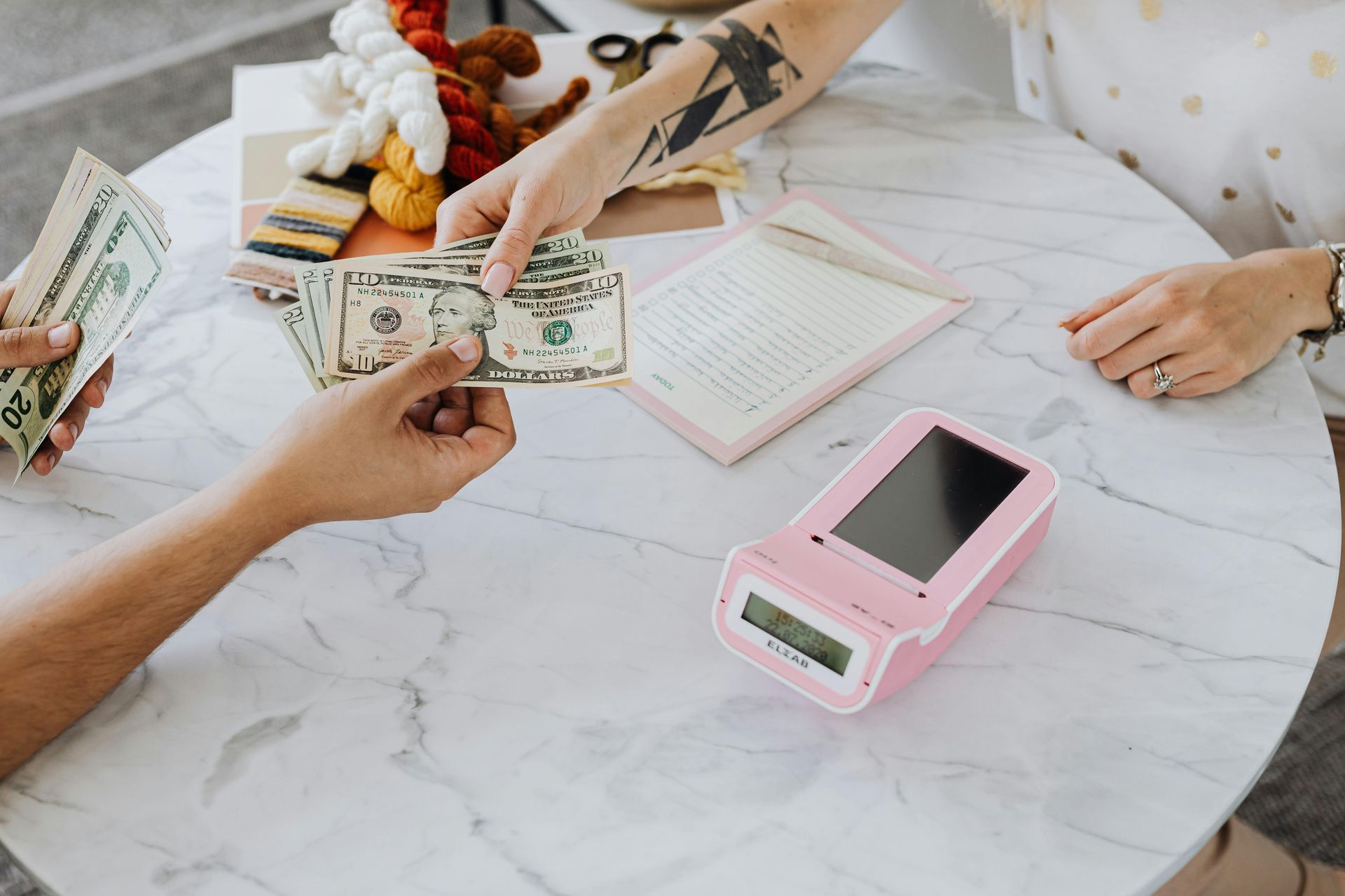 Person handing cash to another person, payment transaction with a pink card reader on a table.