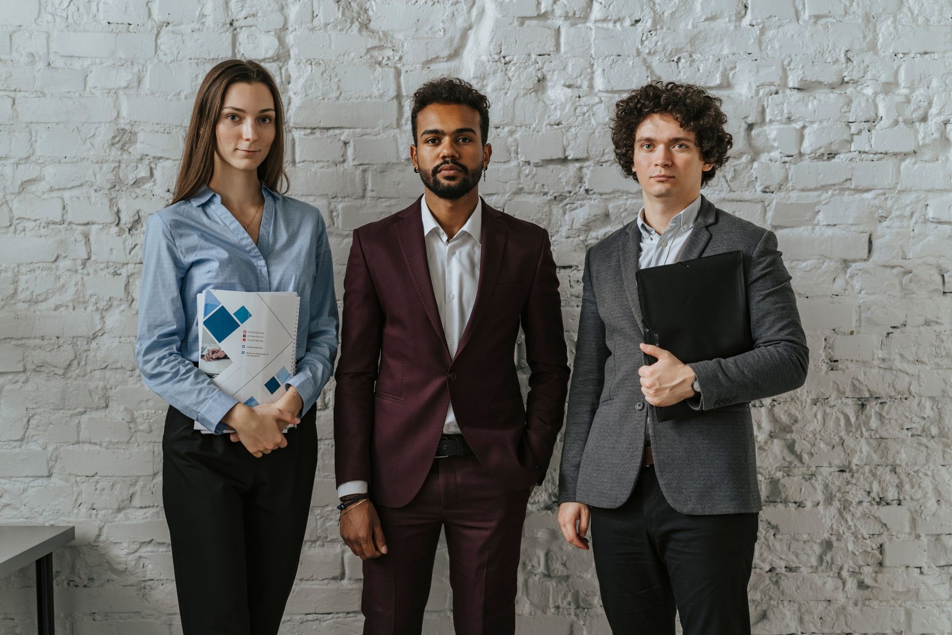Three professionals standing against a white brick wall, one holding a folder, all in business attire.