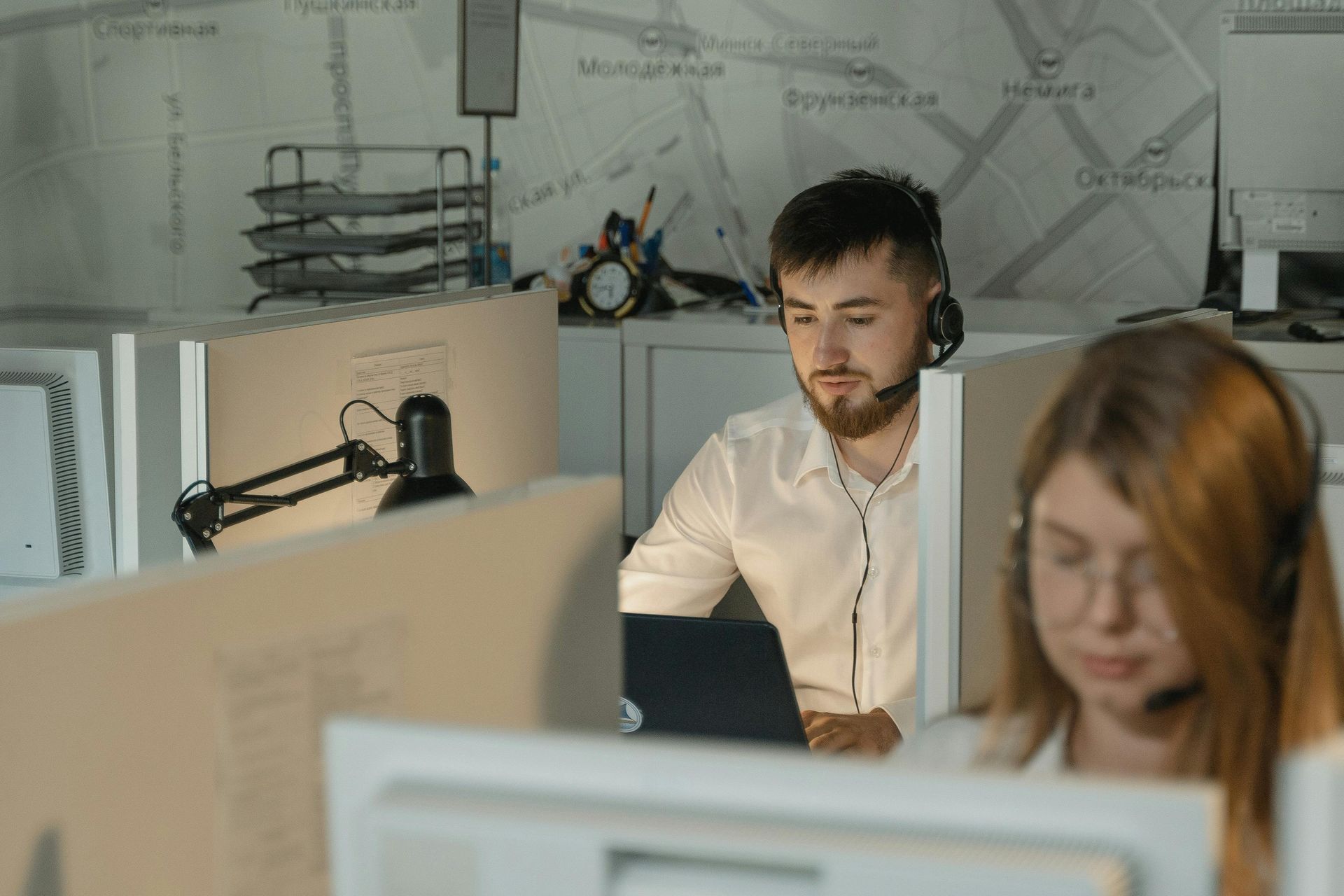 Office workers with headsets at computers, possibly a call center; map on wall in background.