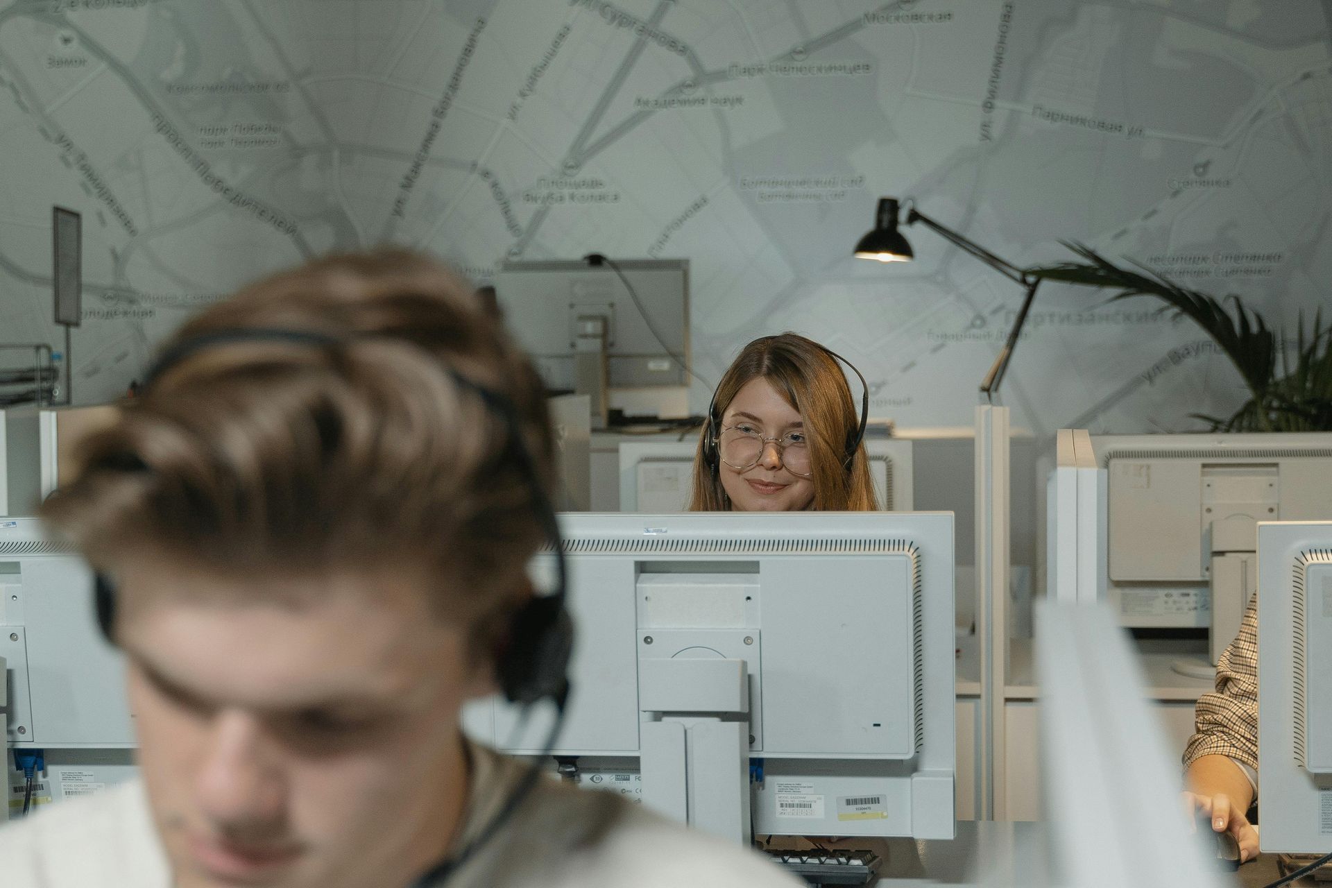 Call center employees wearing headsets work at computers in an office with a map pattern on the wall.