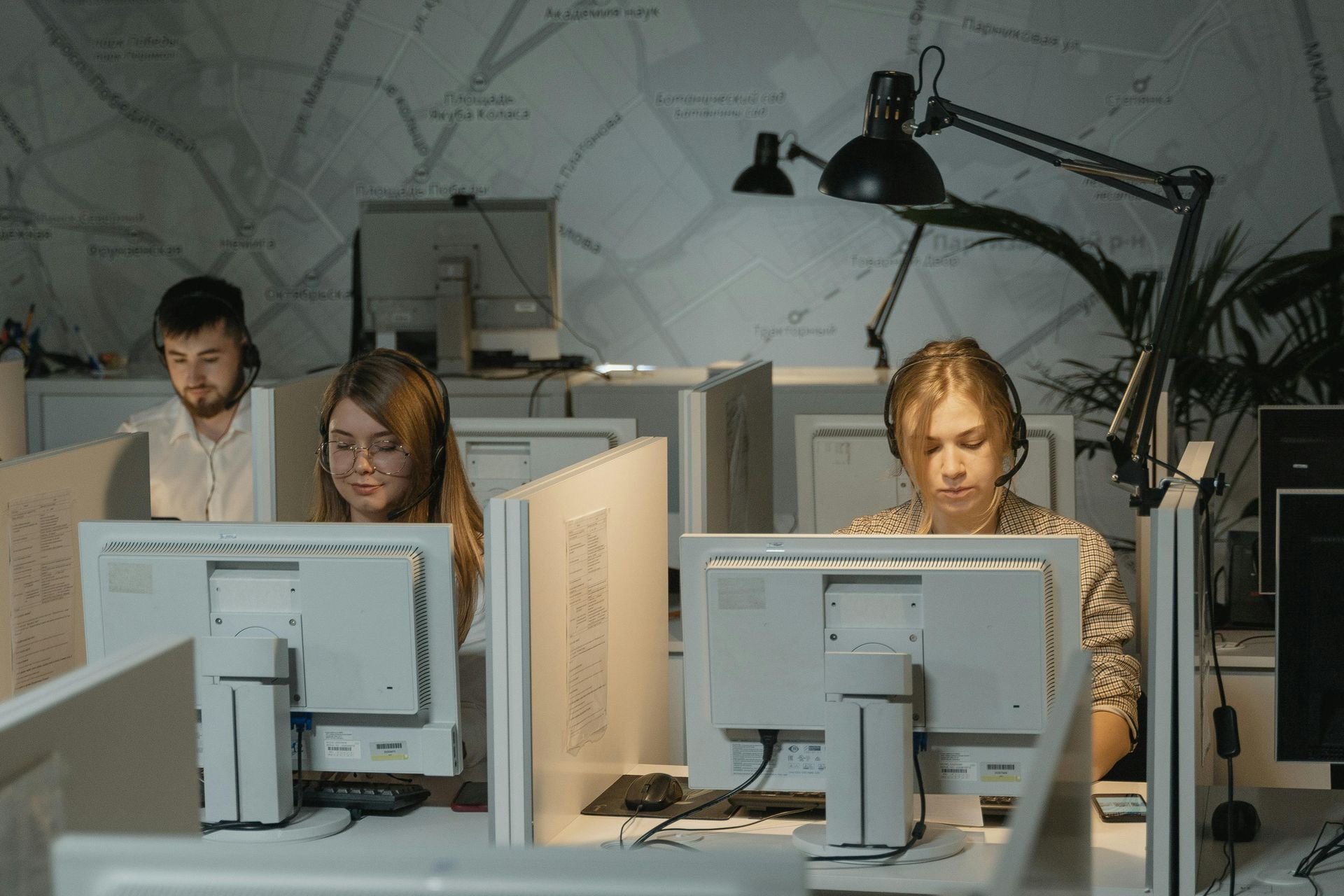 Three people wearing headsets work at computers in an office cubicle setting.