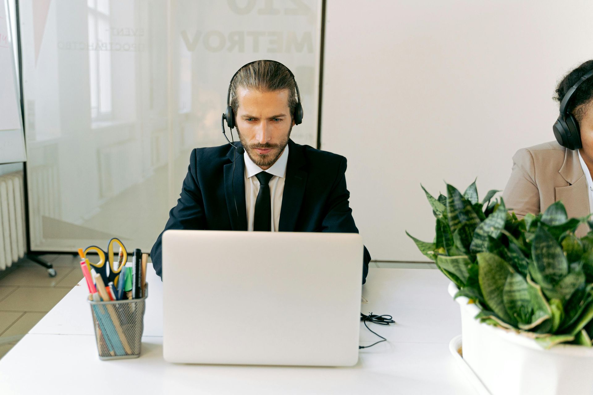 A professional in a suit and headset working on a laptop at a desk with a container of pens and a potted plant nearby.