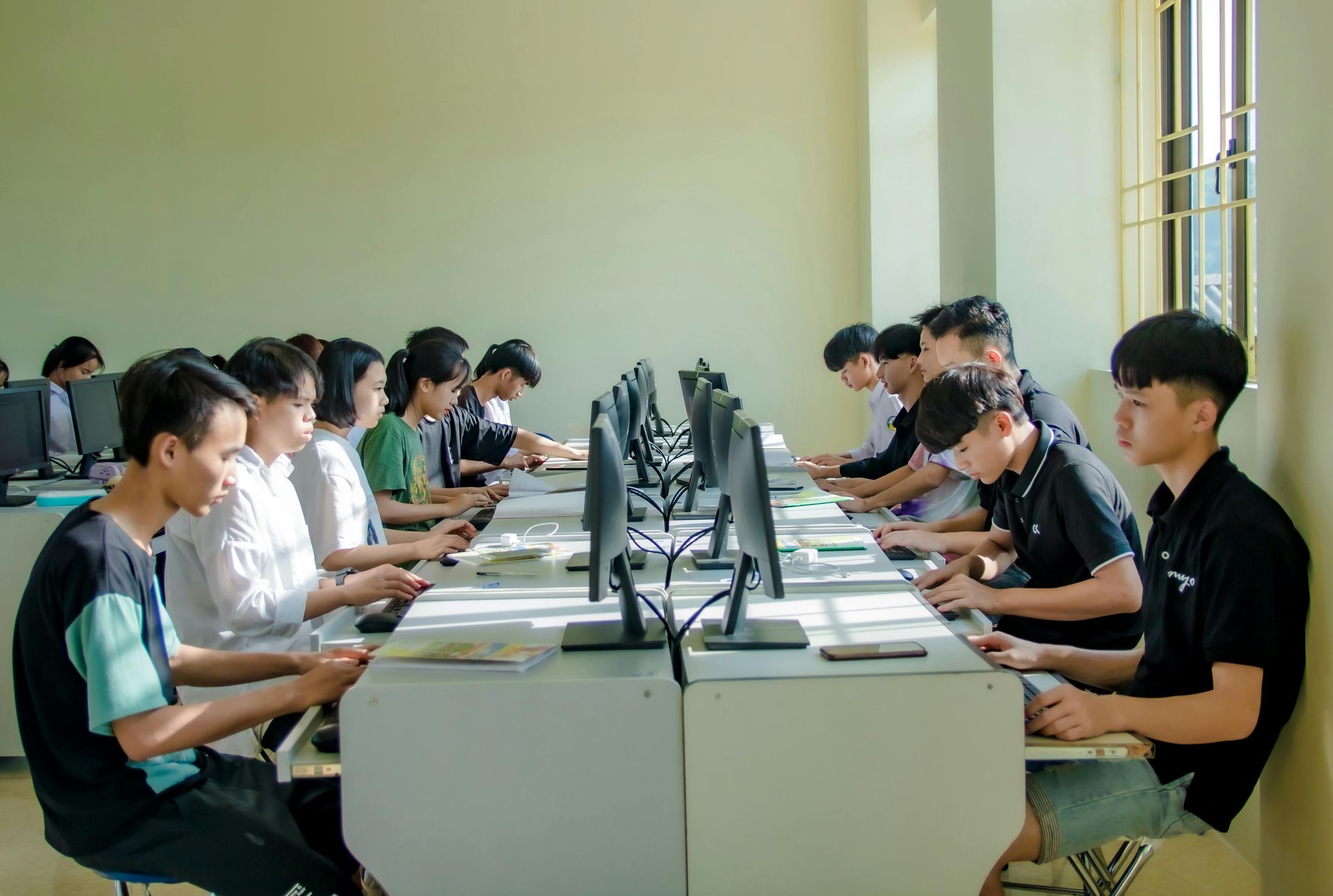 Students sit at computers in a classroom, engaged in study or work at their individual desks.