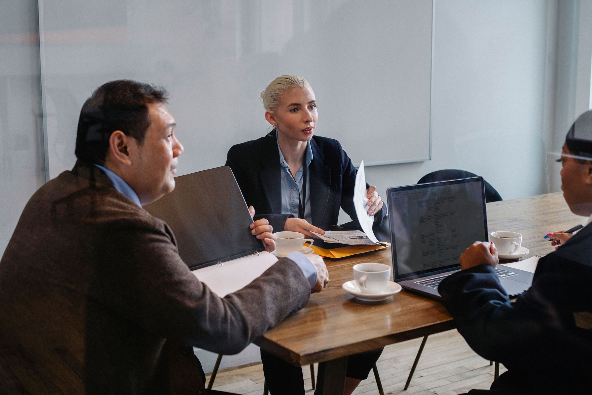 Three people in a meeting around a wooden table, discussing documents in a bright office.