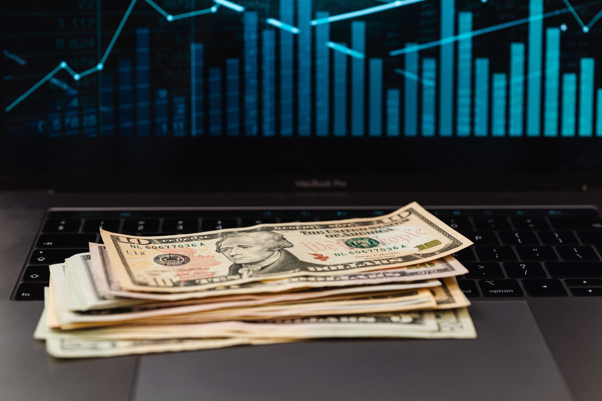 Stack of dollar bills on a laptop keyboard with a blue financial chart on the screen