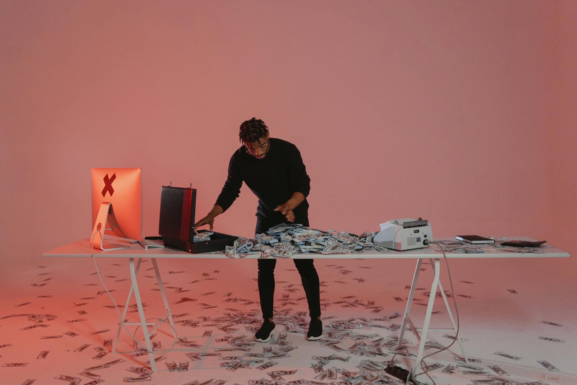 Man surrounded by money, working at a computer on a table in a pink-lit room.