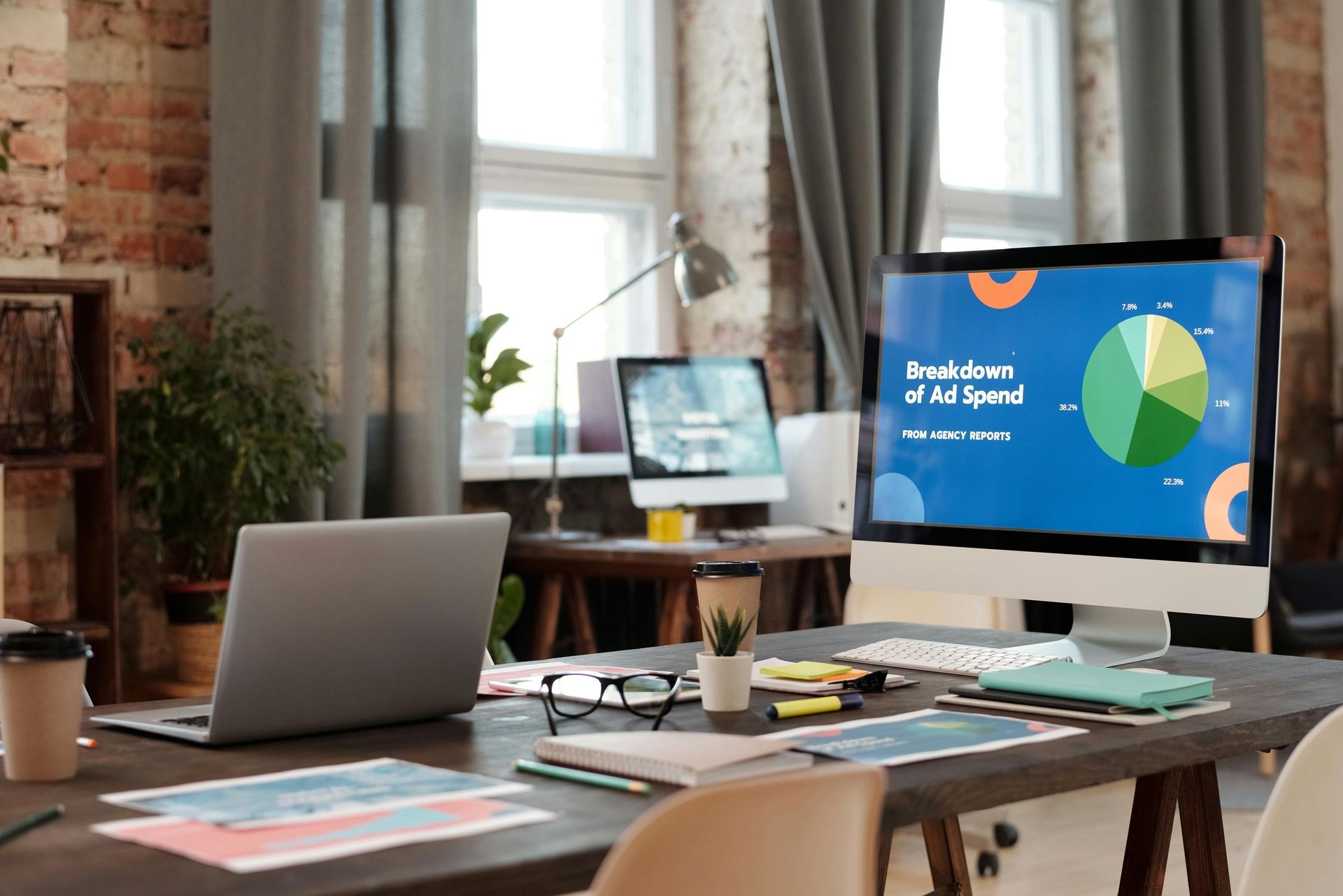 A home office workspace with a laptop, notebooks, coffee, and a monitor displaying a pie chart titled