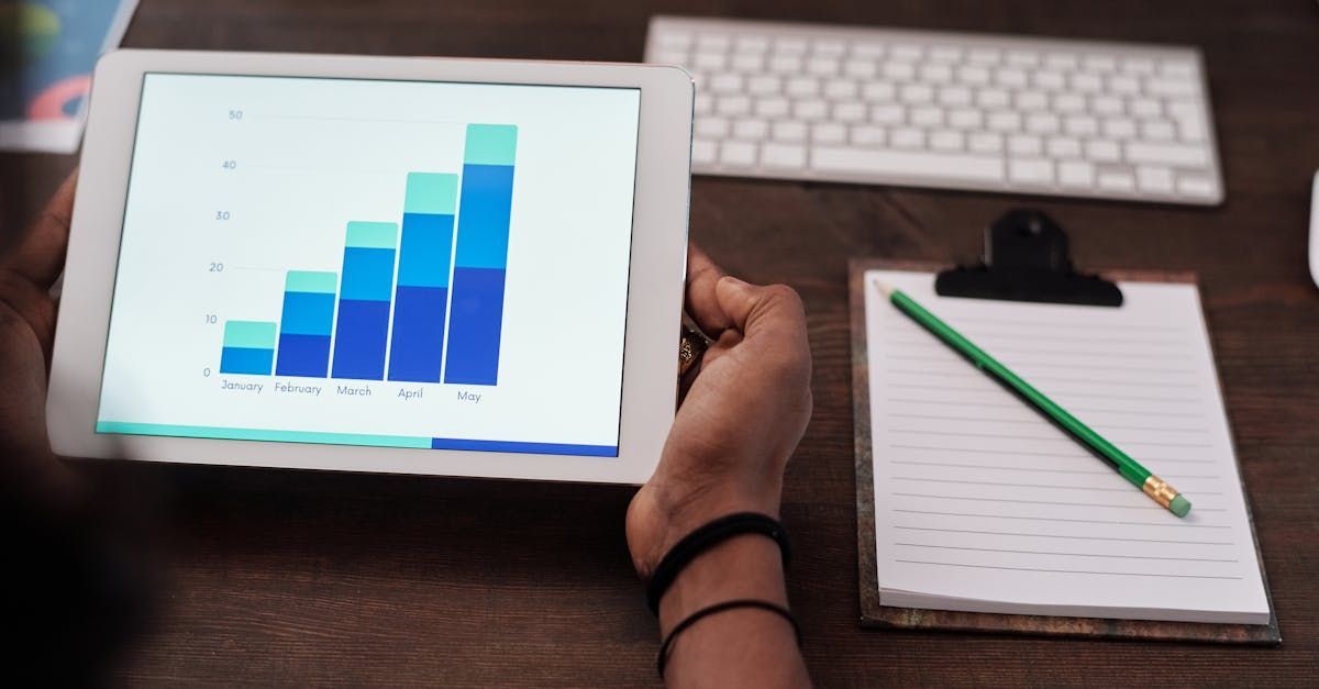 A person holds a tablet displaying a rising bar chart, with a notepad, pencil, and keyboard on a desk nearby.