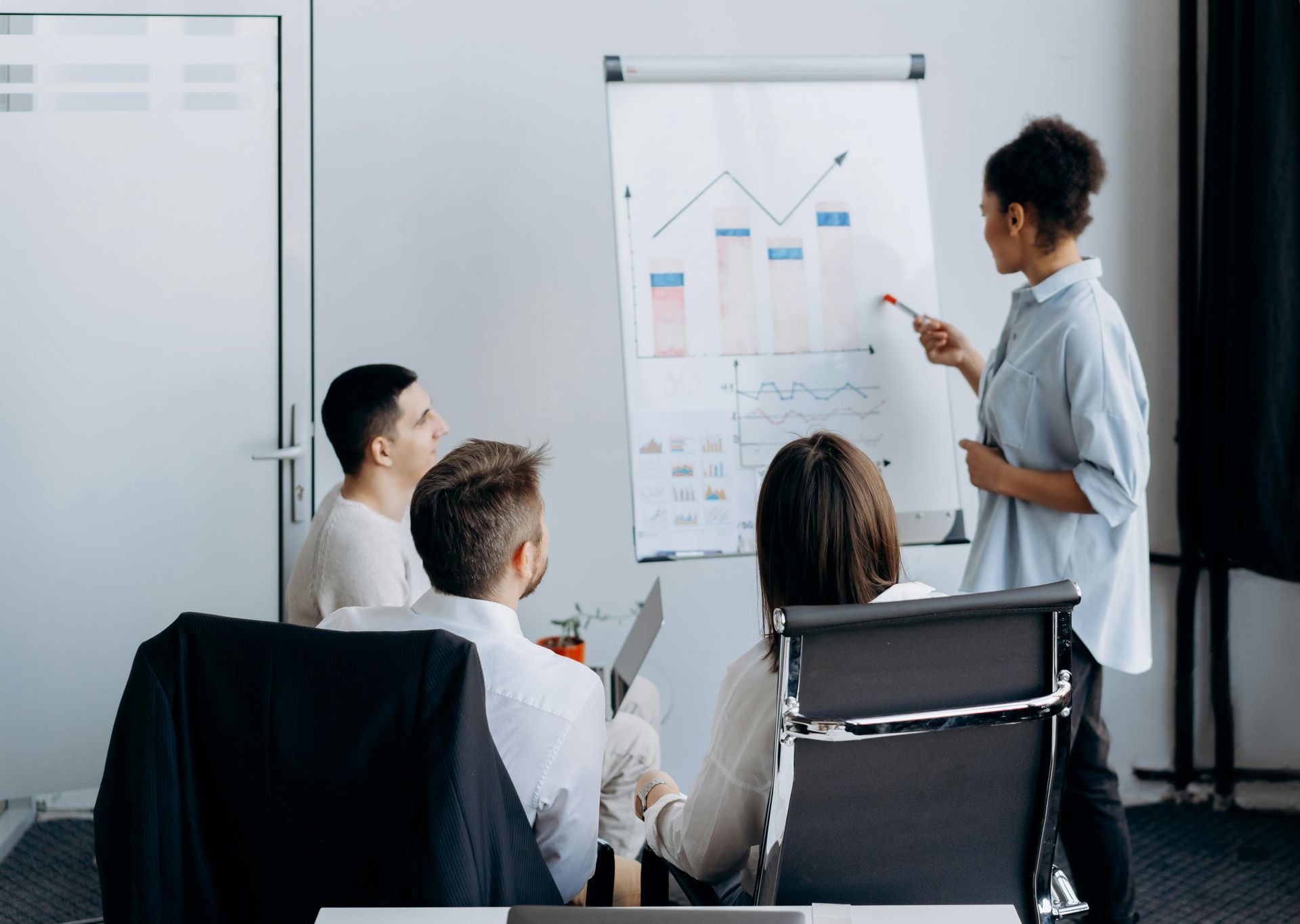 A presenter points to a bar chart on a whiteboard while three colleagues listen during an office meeting.
