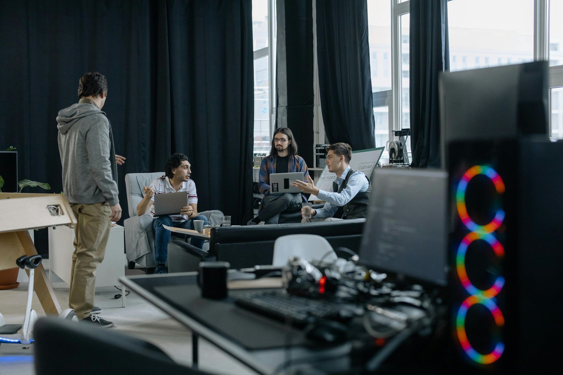 Four people collaborate in a modern, open-plan office, with a foreground focus on a desktop PC featuring RGB lighting.