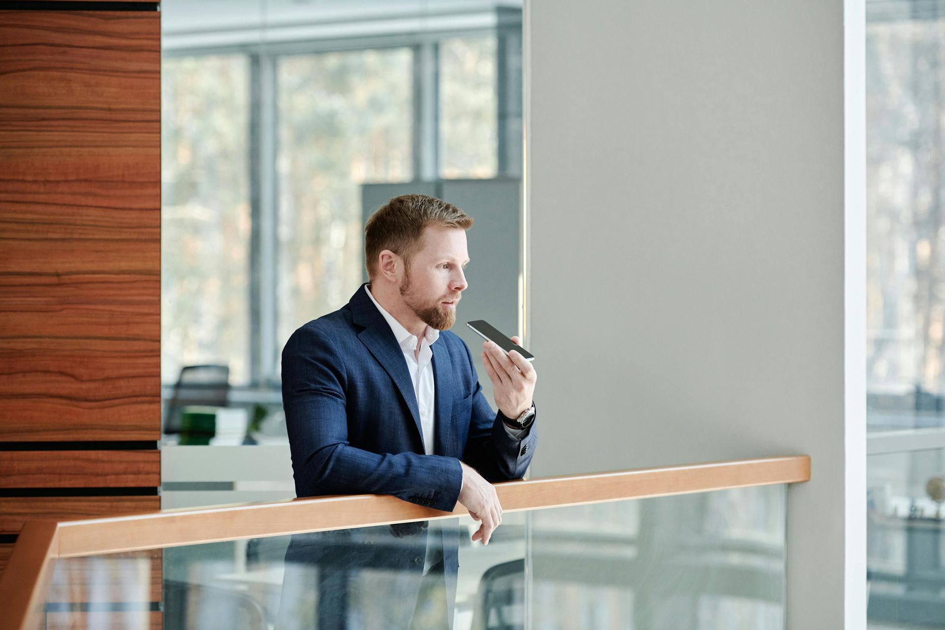 A person in a blue suit leans on a railing in a modern office, holding a smartphone to their mouth to record a message.