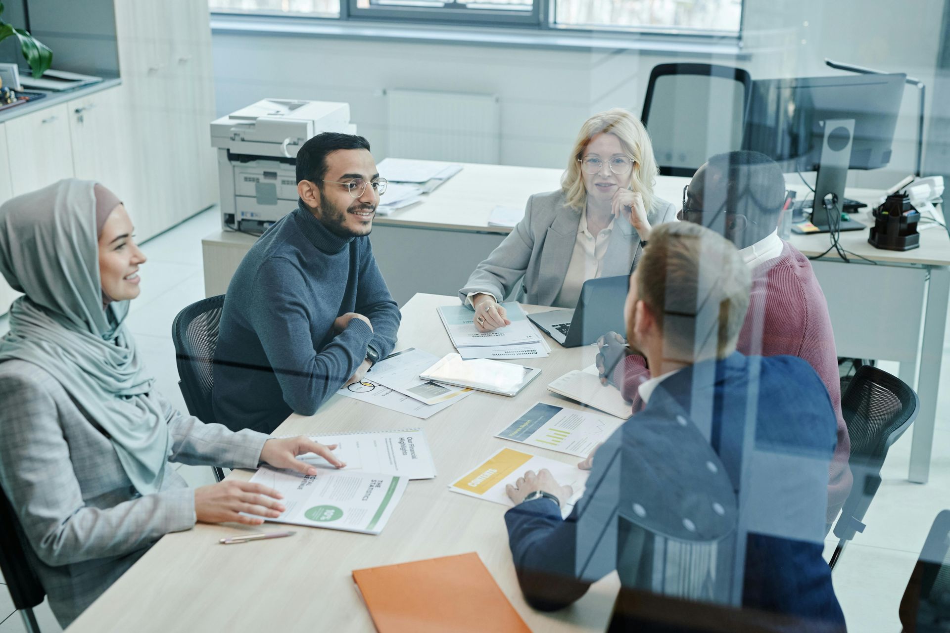 Five professionals sit around a bright office table, reviewing documents and discussing work during a team meeting.