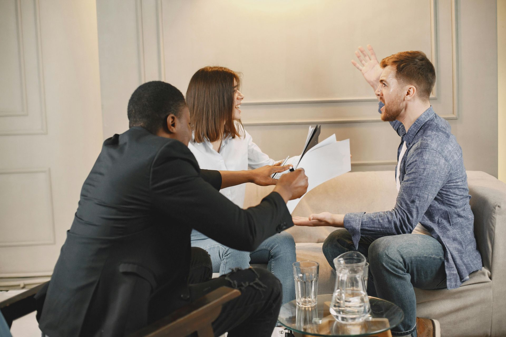 Three people in a meeting room, one gesturing while looking at documents held by another.