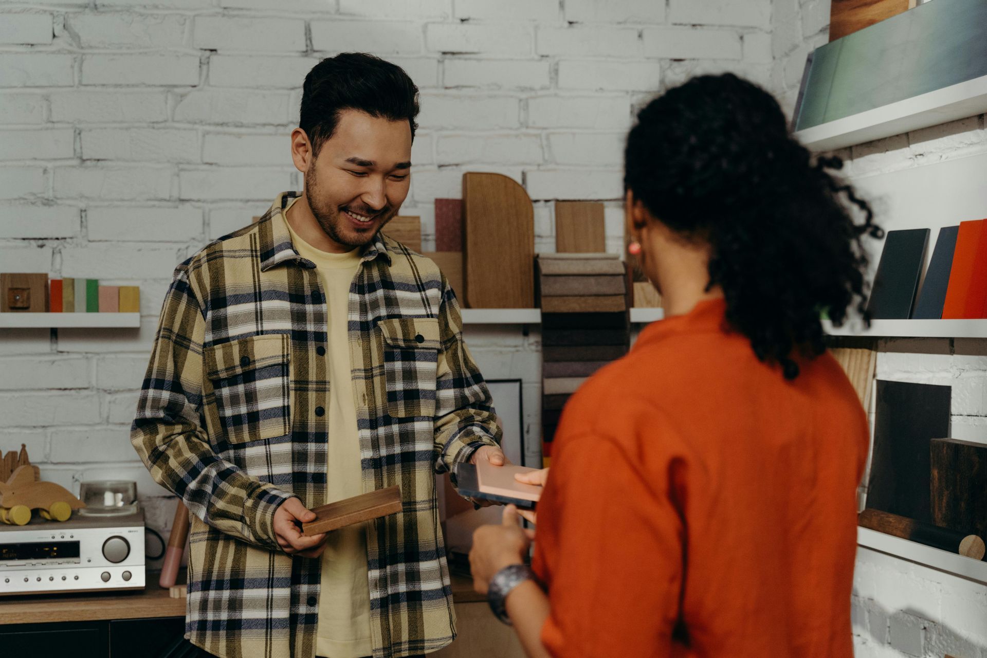 Man shows a wood sample to a person in an orange shirt. They smile in a workshop with shelves.