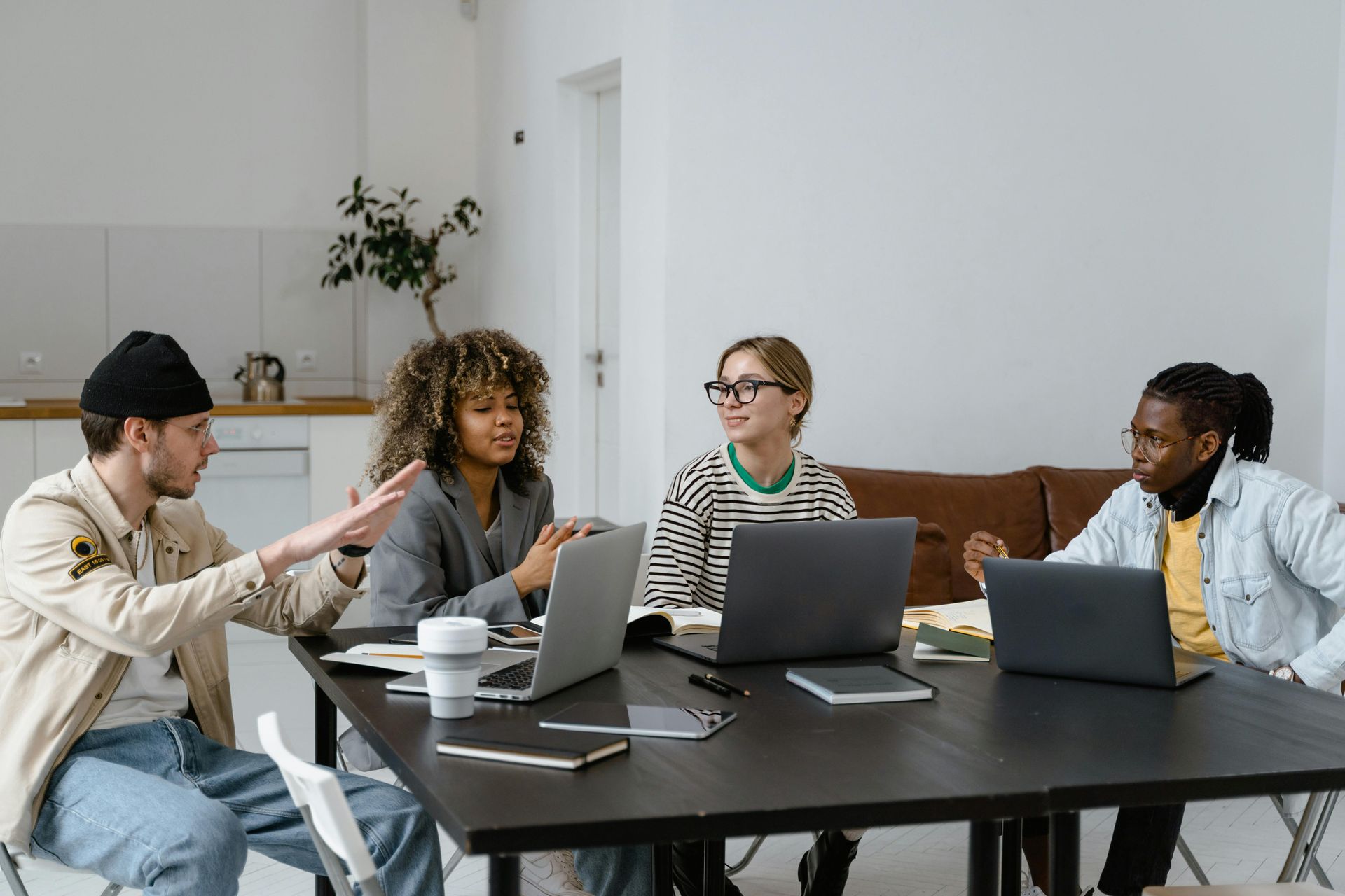 Four people sit around a table in a bright office, working on laptops and discussing a project.
