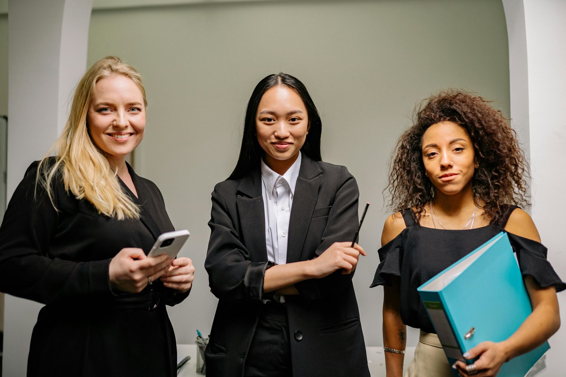 Three professionals stand in an office, one holding a phone, one with a pen, and one holding a blue binder.